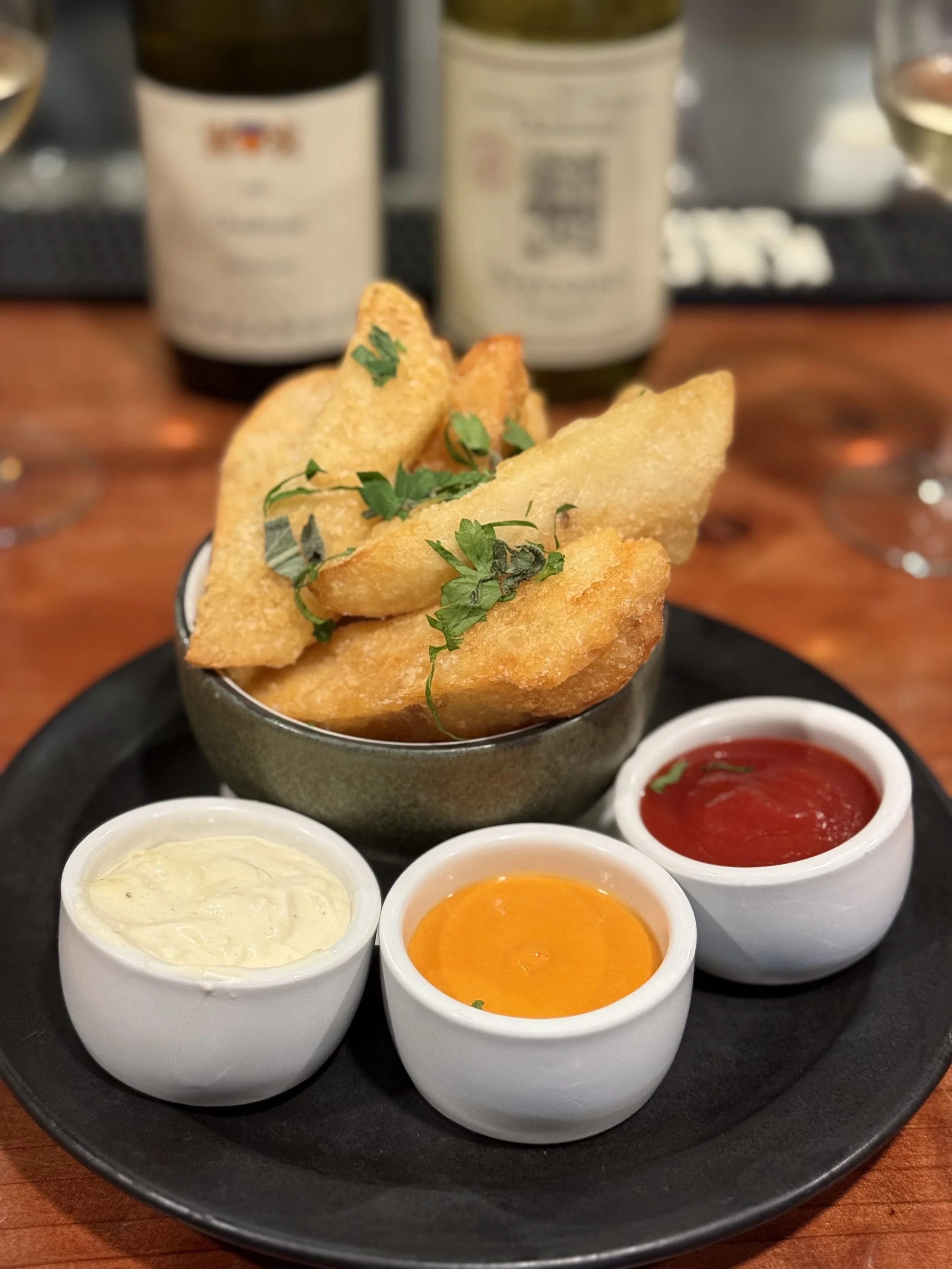 Bowl of fried potato wedges garnished with herbs, served with three dipping sauces: mayonnaise, hot sauce, and yellow mustard or cheese sauce, on a black plate on a wooden table.