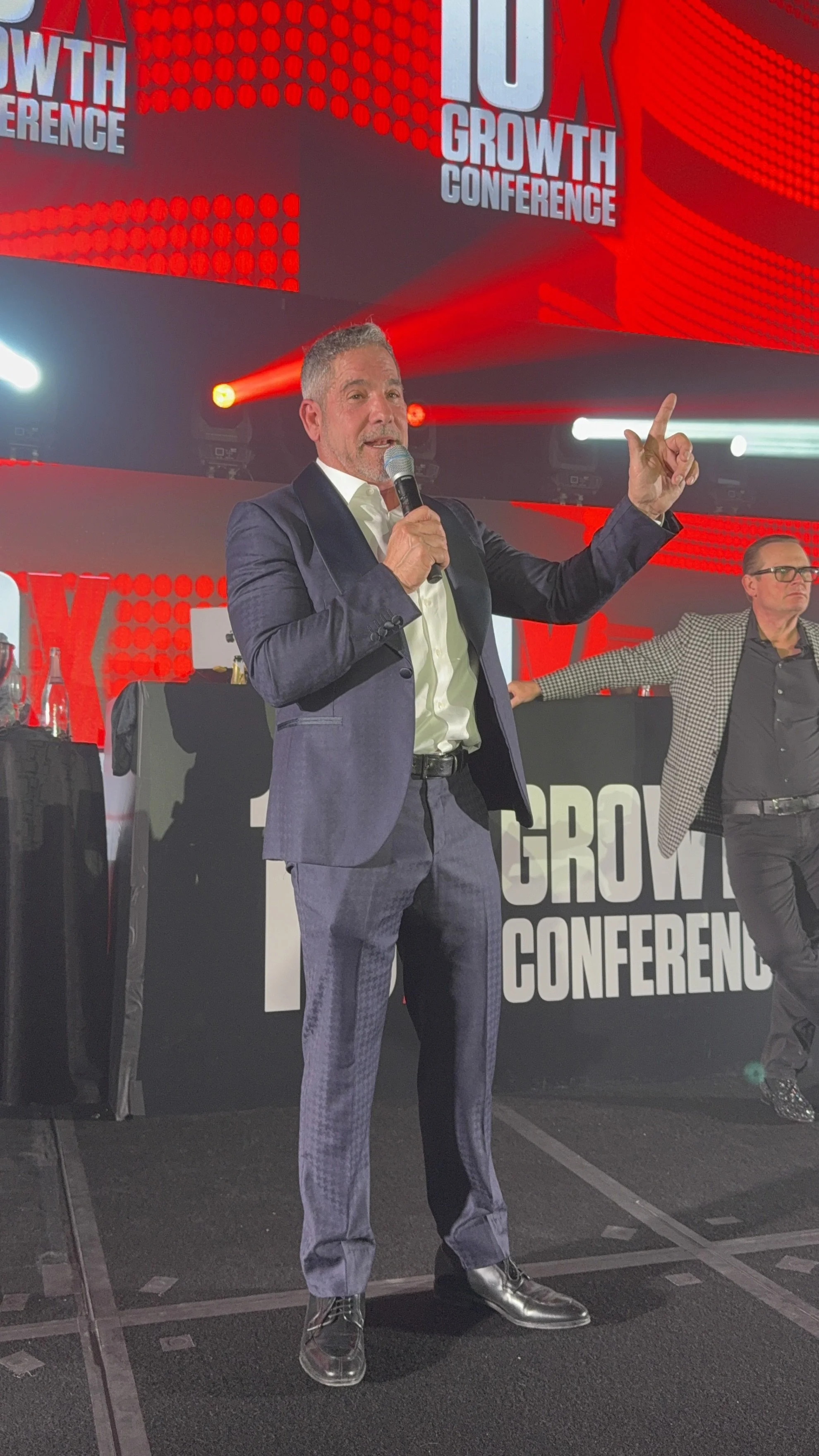 A man in a suit holding a microphone speaking on stage at a conference with the sign '10X Growth Conference' in the background.