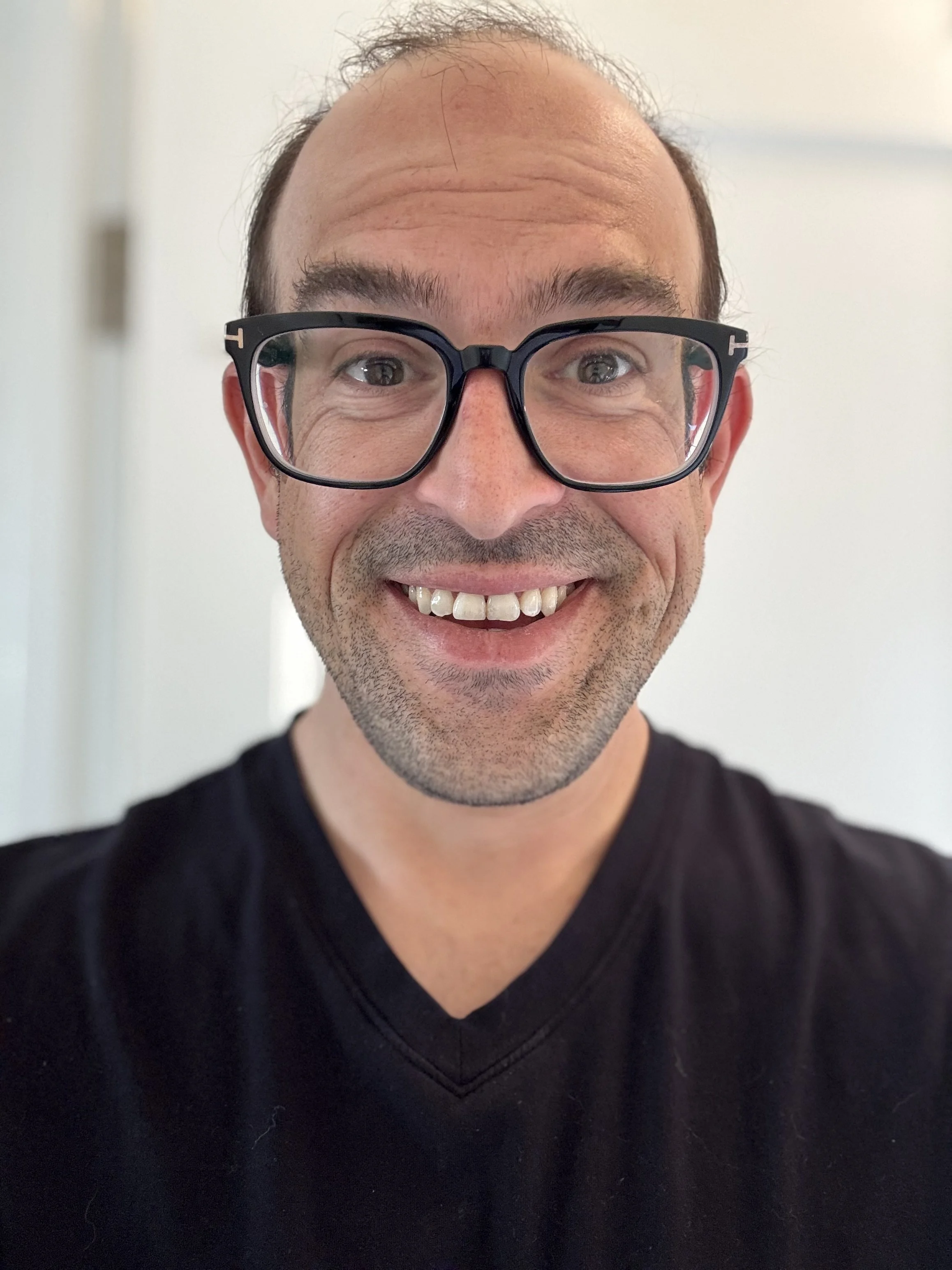 A man with glasses smiling at the camera, wearing a black shirt, against a plain background.