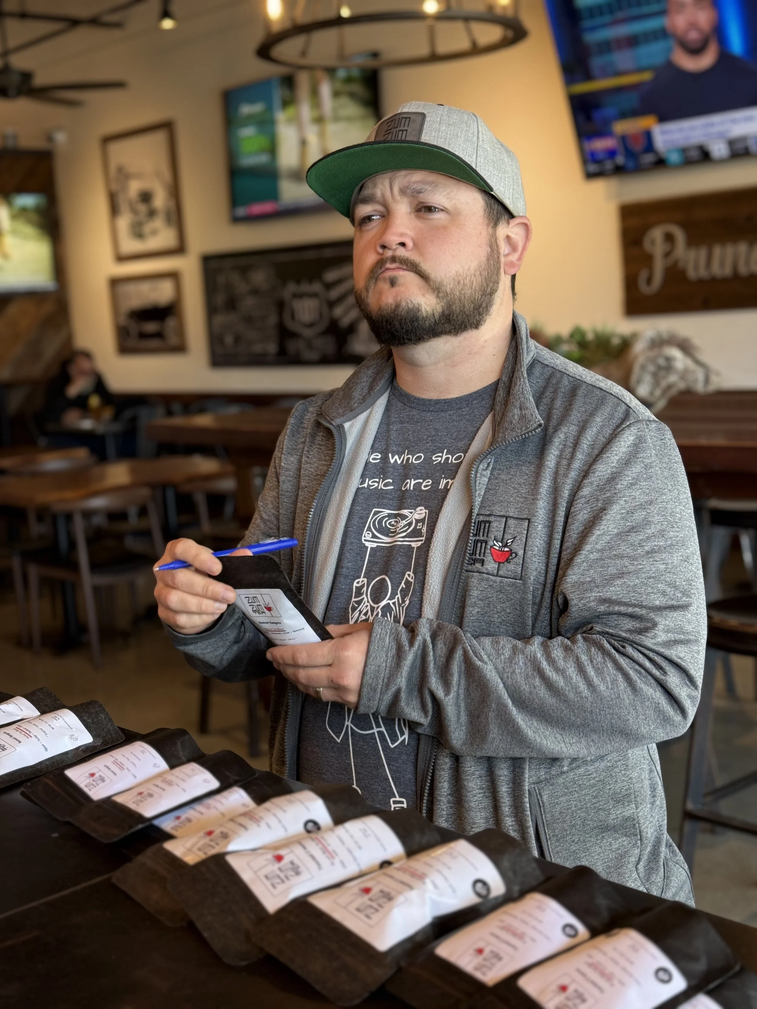A man with a beard and gray cap tasting coffee at a restaurant with several coffee packets on the table.
