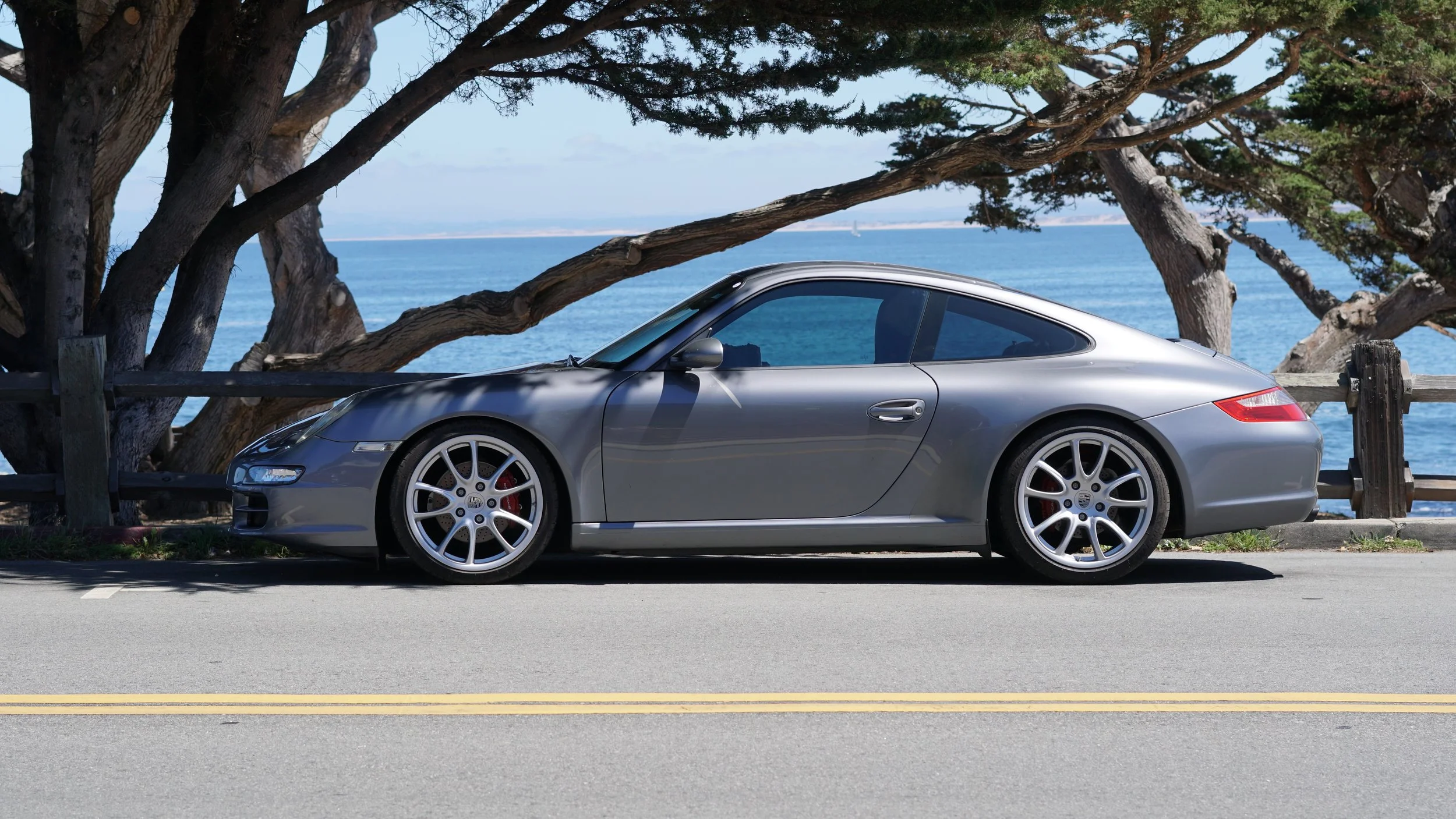 A silver Porsche sports car parked beside a wooden fence by the ocean, with trees and blue sky in the background.