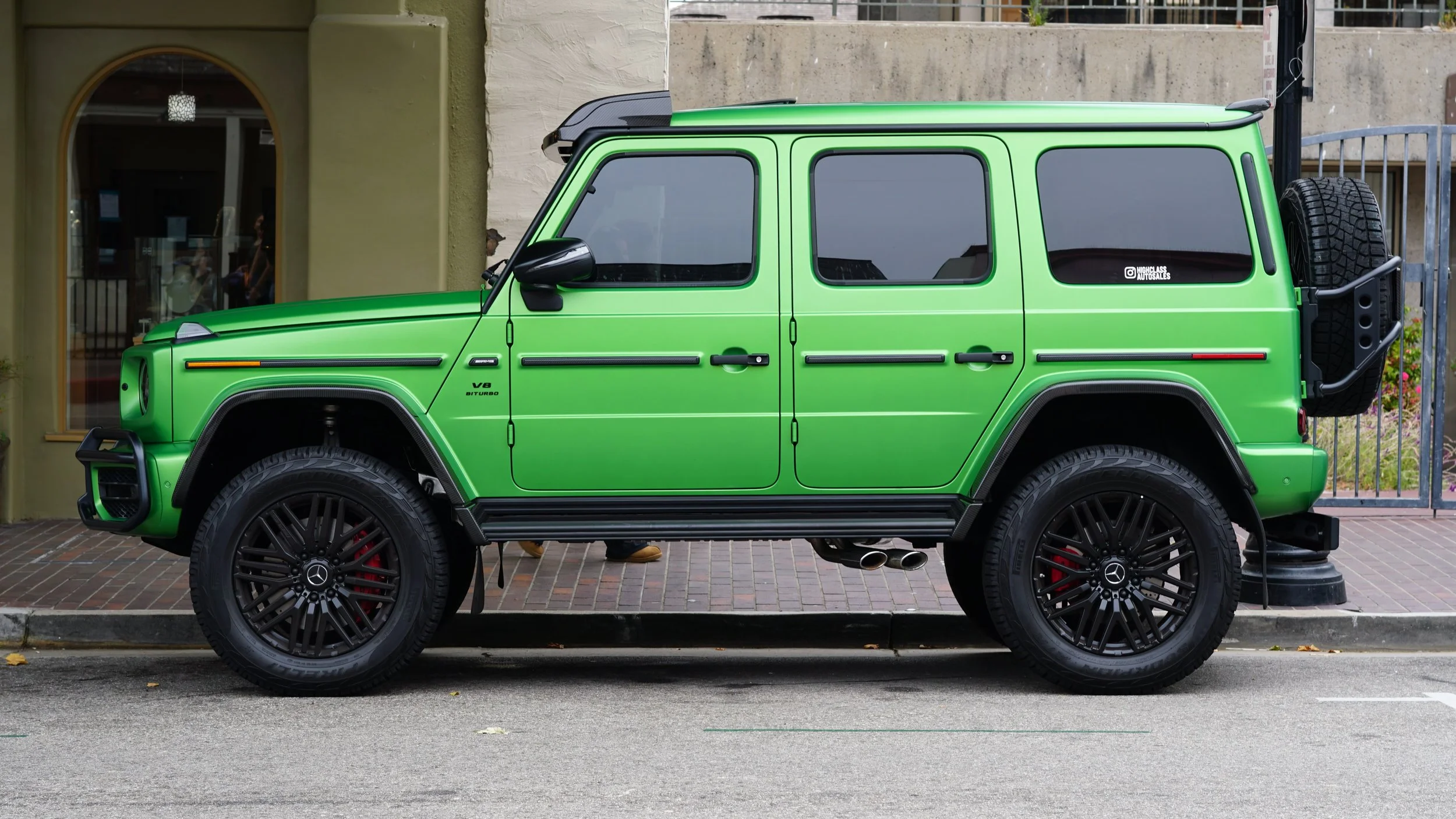 Green Mercedes-Benz G-Class SUV parked on the street in front of a building with large windows and a gate, featuring black wheels, a spare tire at the back, and a sticker on the rear window.
