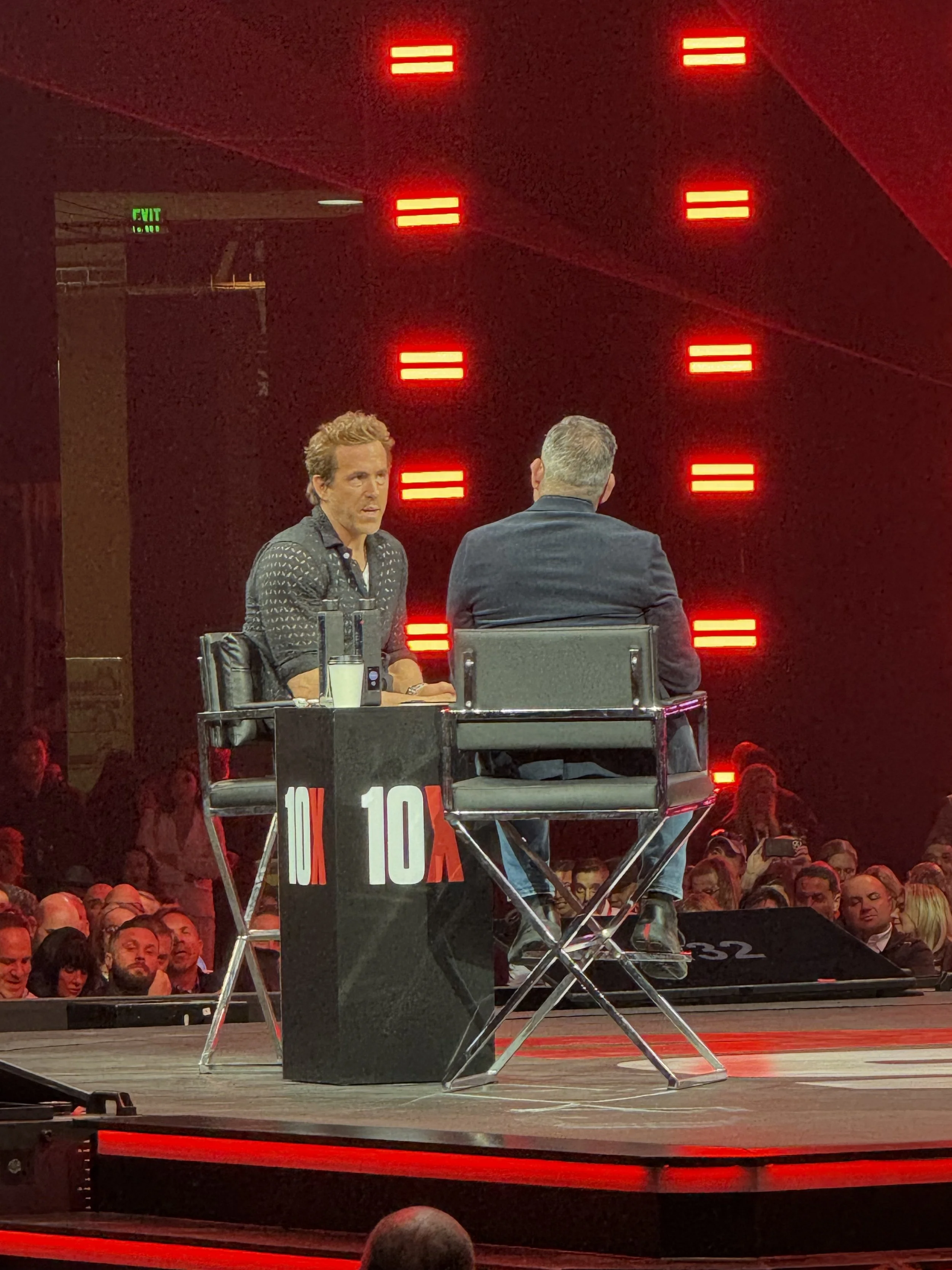 Two men engaged in discussion on stage during a panel at a live event, with audience watching, red illuminated wall background, and a table labeled '10A' between them.