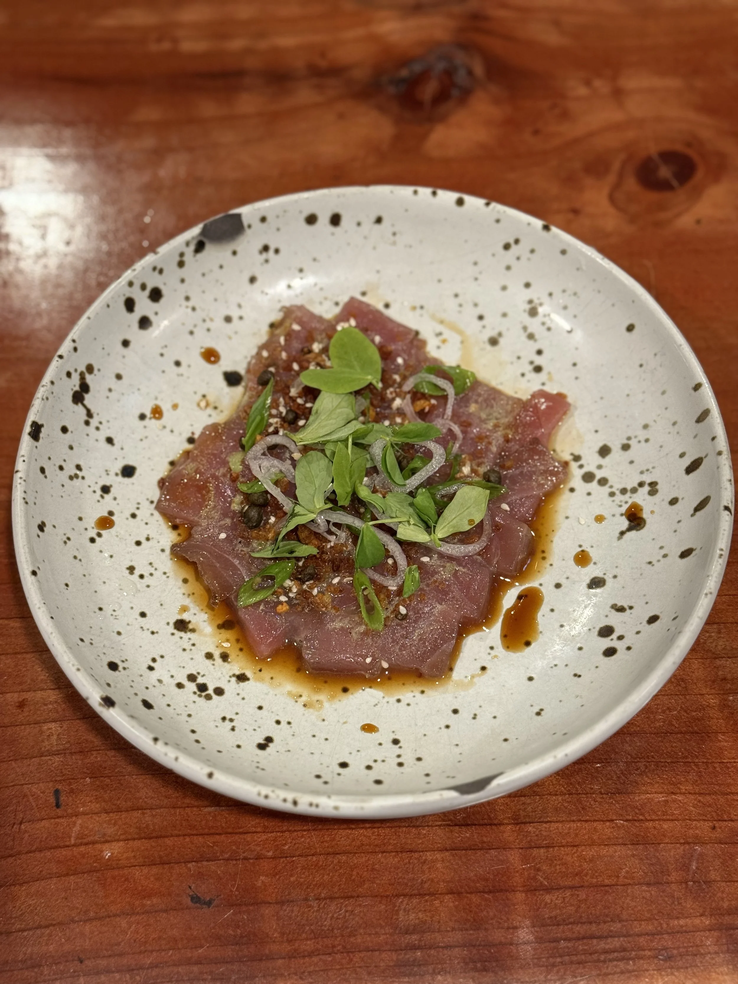 Slices of raw tuna sashimi topped with green herbs, sliced onions, black pepper, and sauce on a white speckled plate with a wooden table underneath.