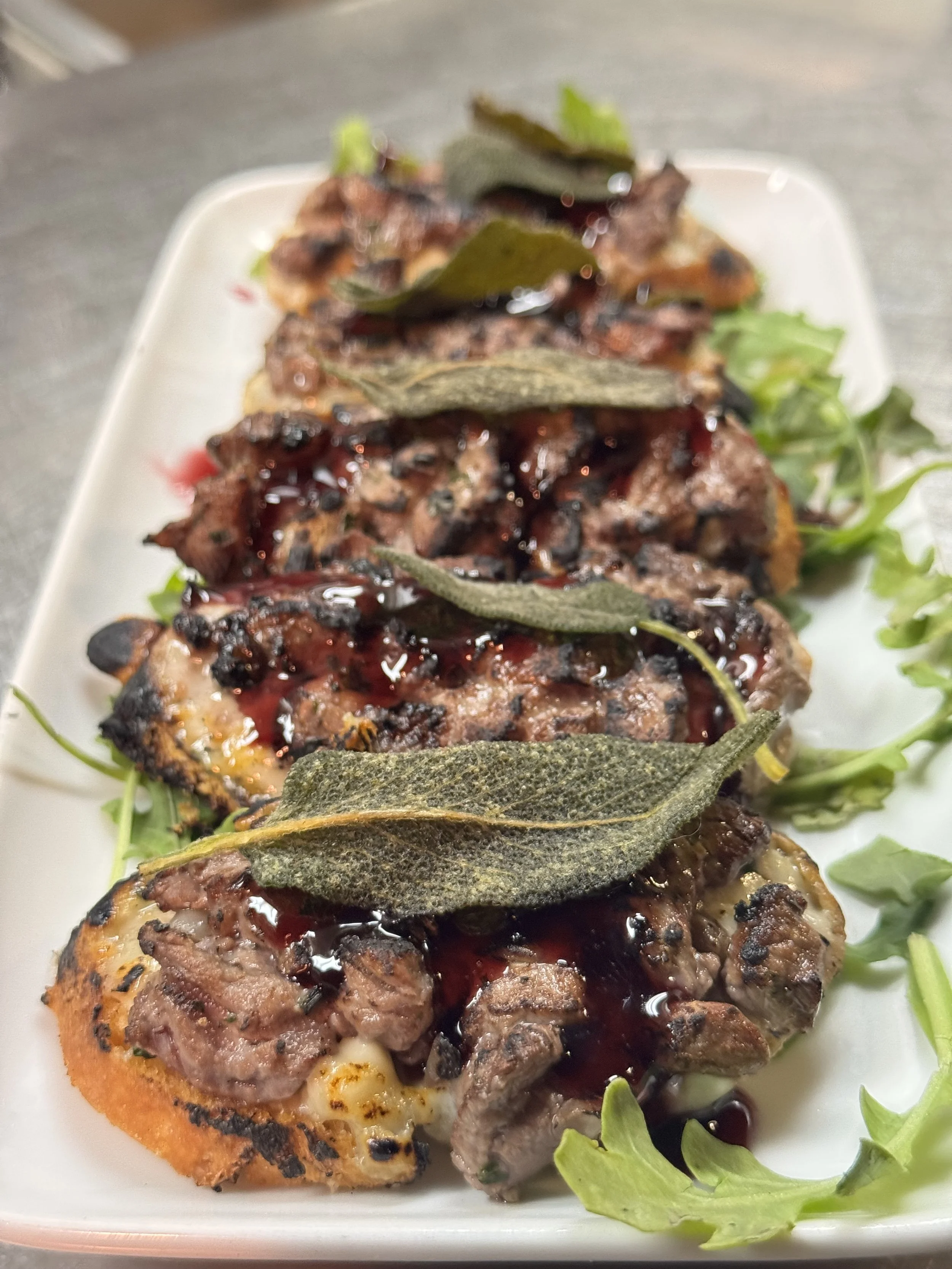 Four grilled meat pieces topped with bay leaves, drizzled with dark sauce, served on a white rectangular plate with green leafy garnish on the side.