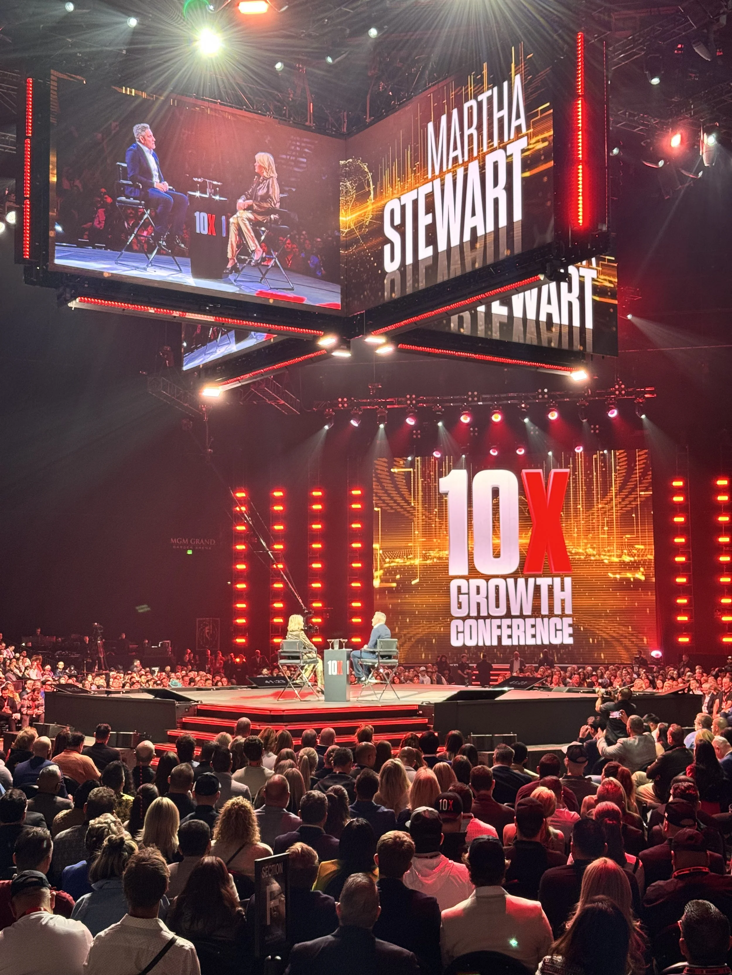 A large conference stage with a big audience, featuring a big screen displaying 'Martha Stewart' and '10X Growth Conference,' with two people seated on stage engaged in a discussion under colorful lighting.