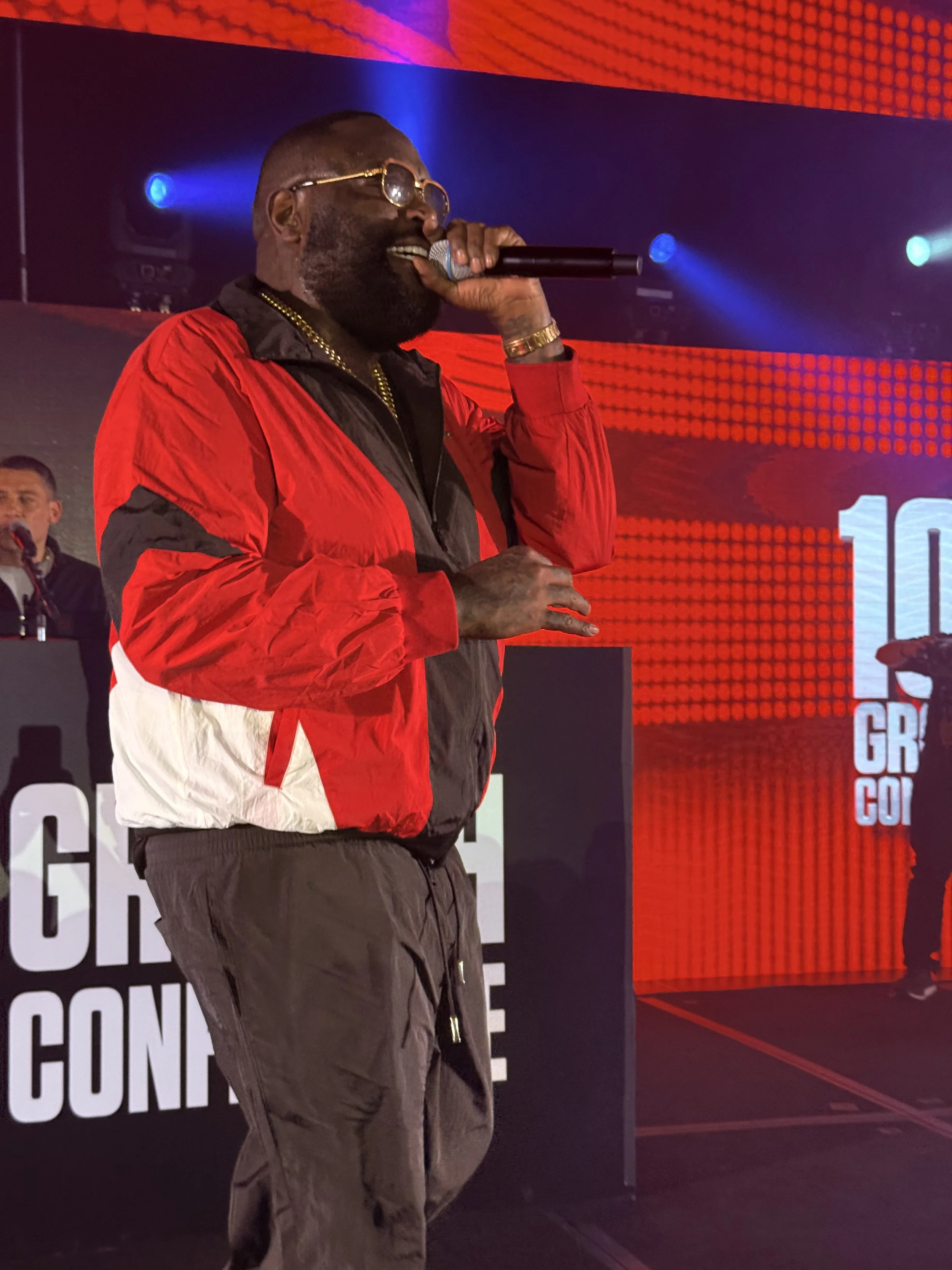 A man wearing sunglasses, a red and black jacket, and dark pants holding a microphone on a stage with a red and black background.