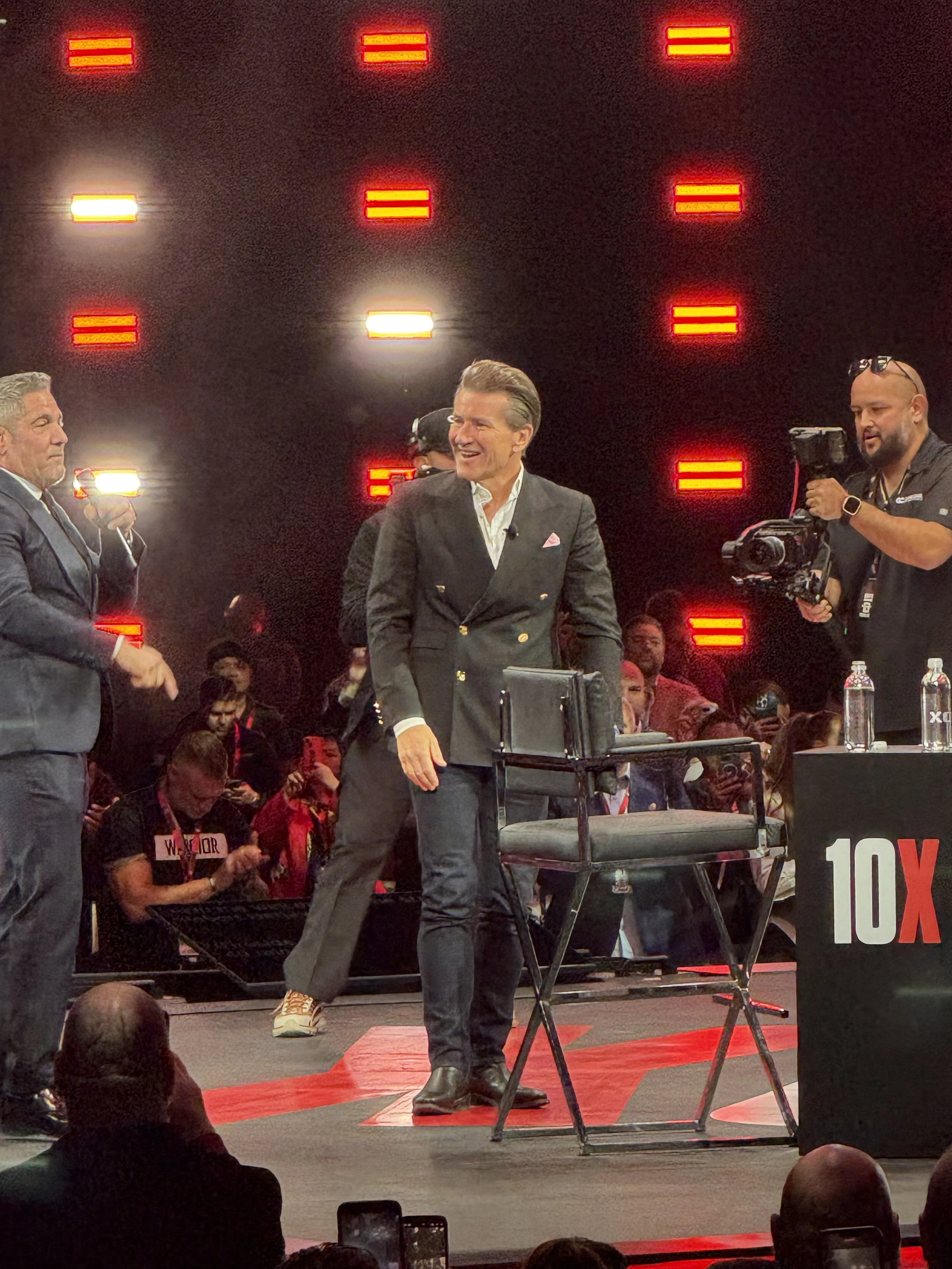 A man in a black double-breasted suit with a white shirt standing on stage, smiling. There are two other men nearby, one in a suit and the other with a camera. The stage background has red illuminated signs, and there's a table with water bottles in