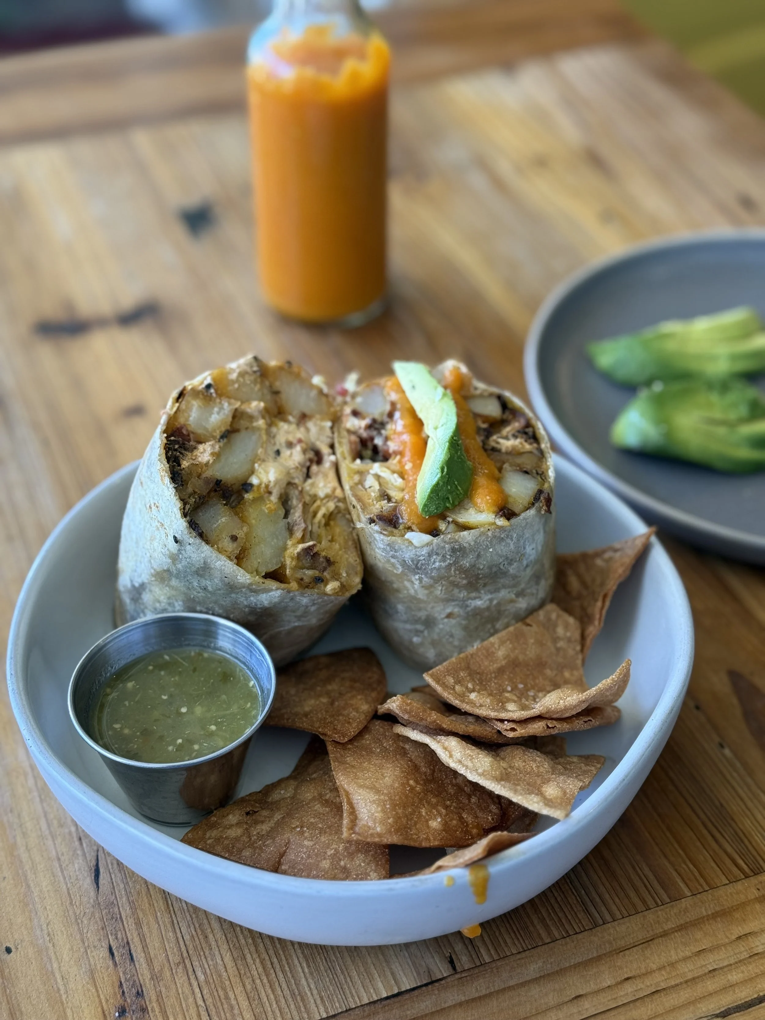 A plate with a burrito cut in half, containing potatoes, cheese, and vegetables, served with a side of chips and a small cup of green salsa. In the background, a bottle of orange sauce and a small bowl of green pickles are visible.