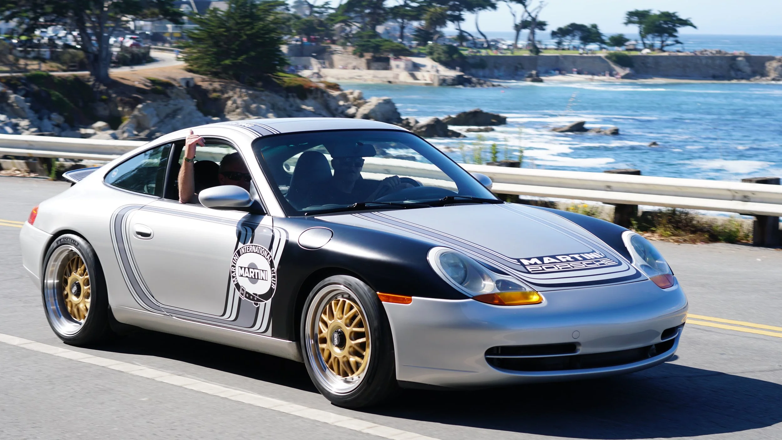A silver Porsche sports car with Martini Racing livery driving along a coastal road with the ocean and trees in the background.