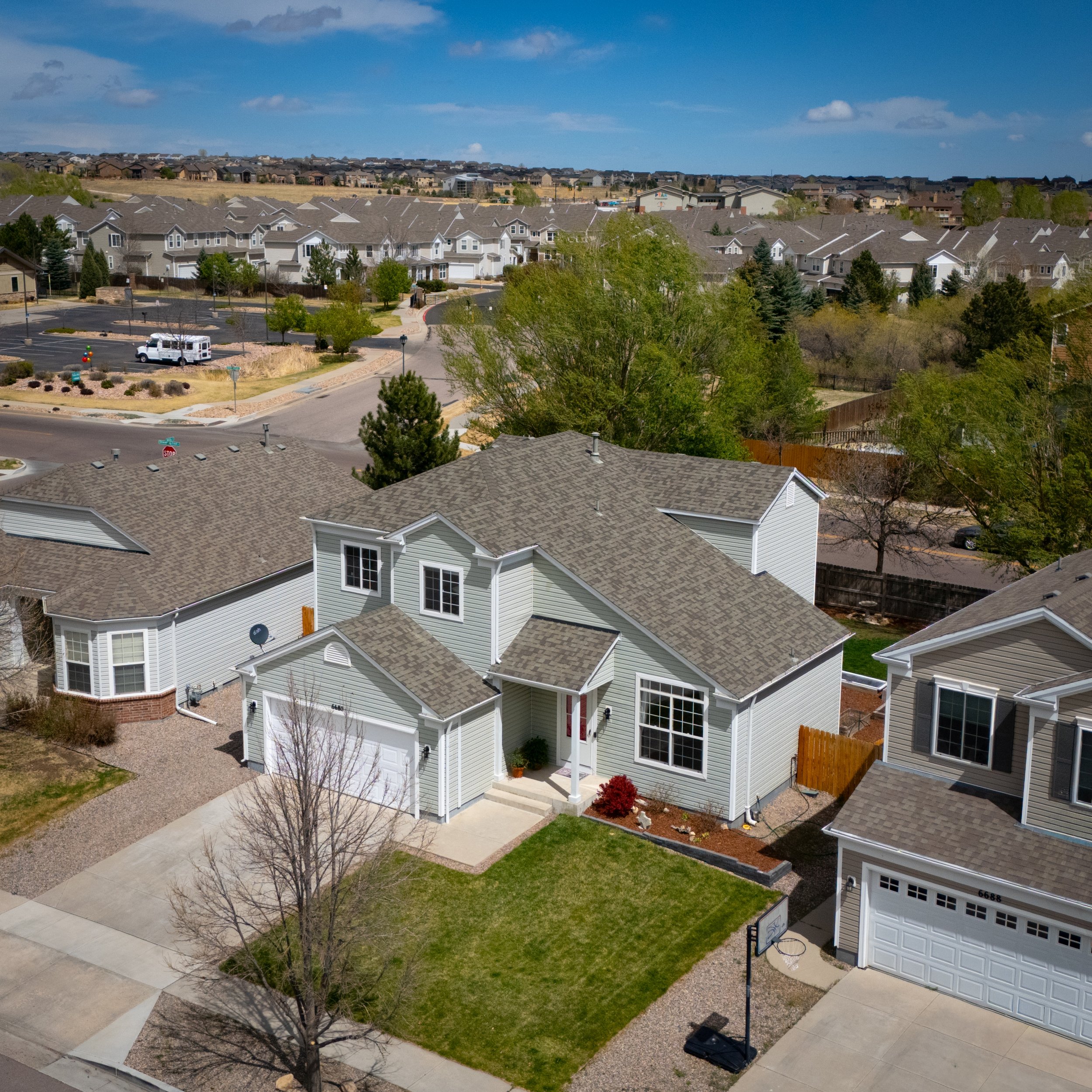 A residential neighborhood with modern houses, green lawns, trees, and a clear blue sky. A small white truck is parked in the driveway, and a basketball hoop is visible in the backyard. The image is taken from an elevated perspective, showing the nei