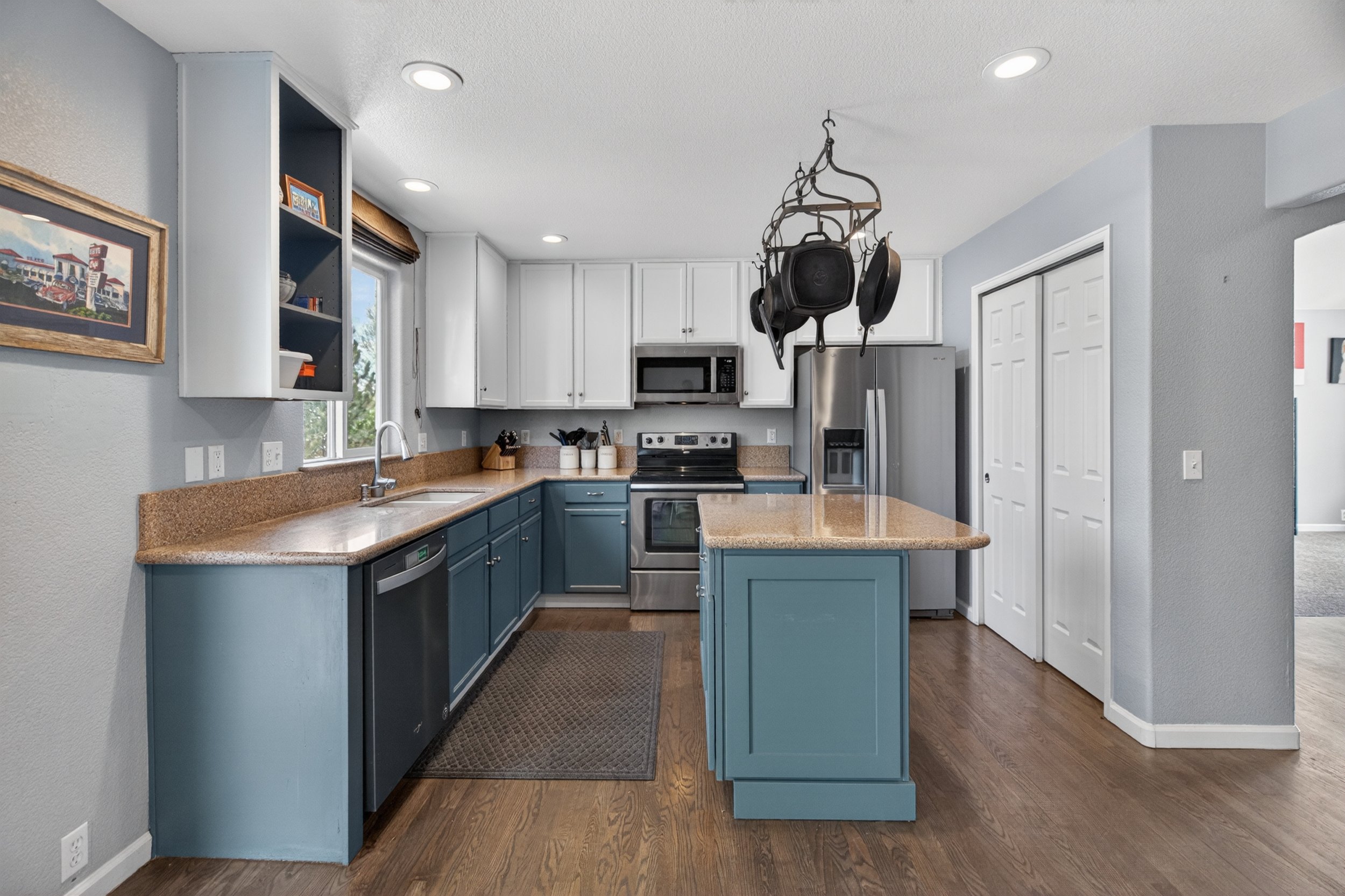 A modern kitchen with blue lower cabinets and white upper cabinets, a beige countertop, stainless steel appliances, and a hanging pot rack with pots and pans, illuminated by recessed ceiling lights.