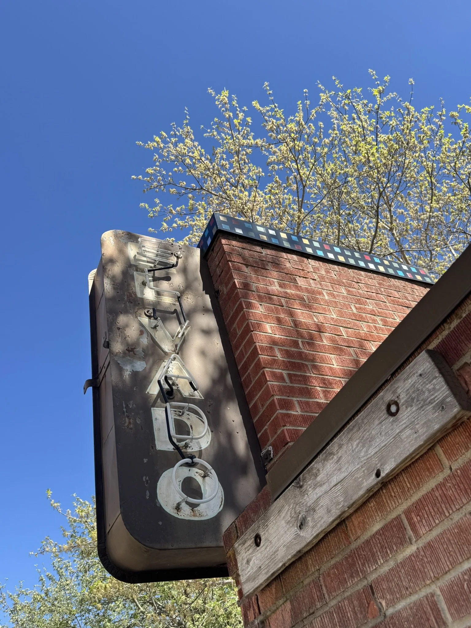 A brick chimney with a decorative tile cap and an outdoor electrical box with multiple wires, mounted on a brick wall, with a clear blue sky and trees in the background.