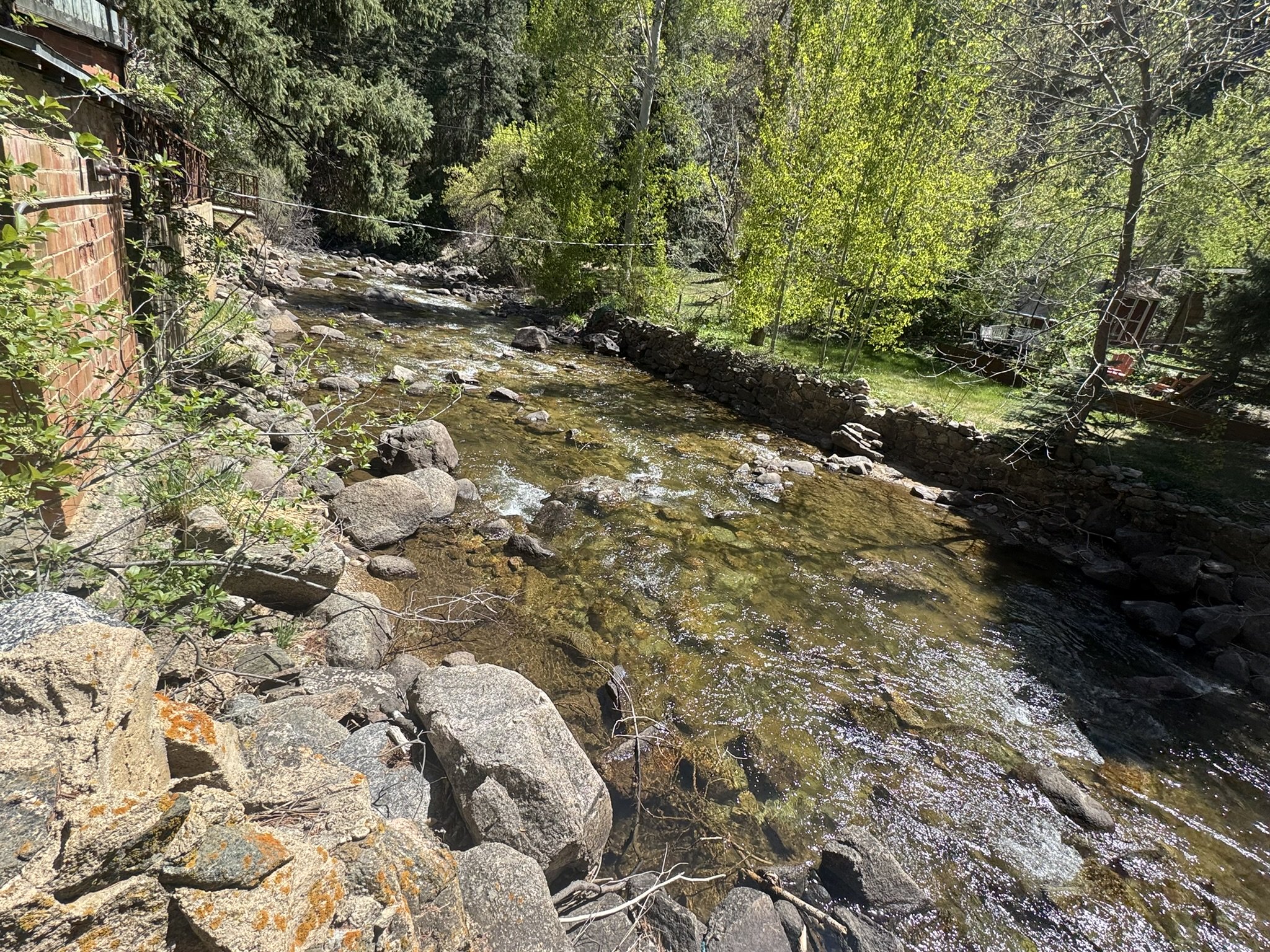 A creek flowing through a wooded area with rocks along the water's edge and a stone wall on the left side. Trees with green leaves line the creek, and a house with a patio is visible in the background.