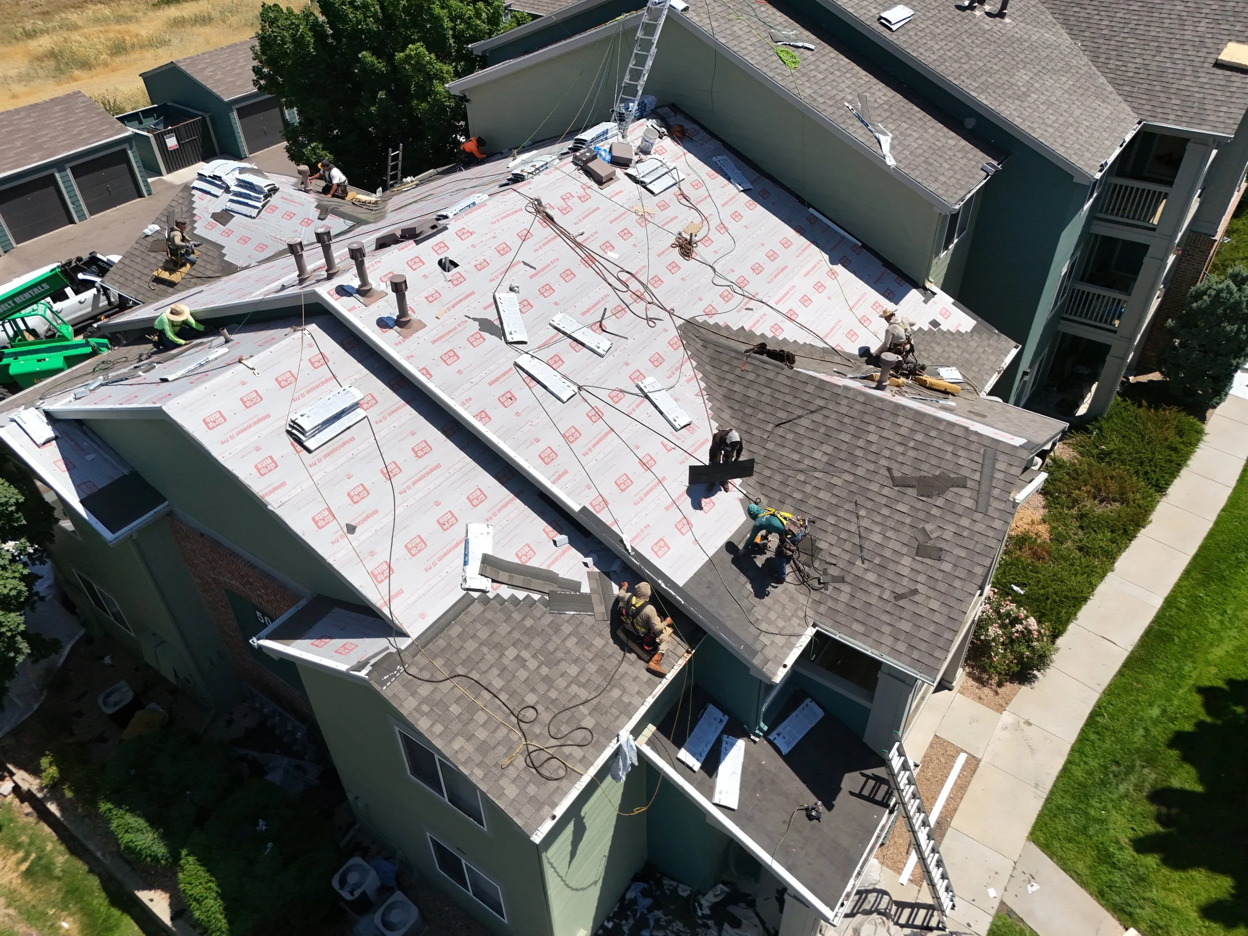 Drone photo of a multi-story residential building under renovation, with workers on the roof installing new roofing materials, surrounded by trees, a sidewalk, and neighboring houses.