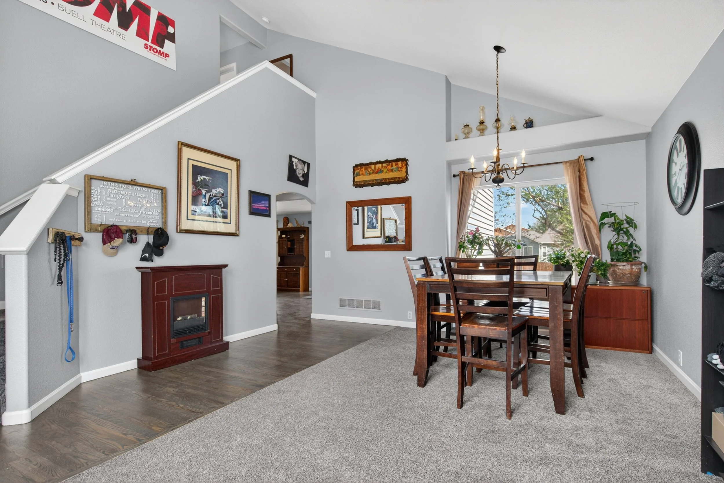 A living room with a dining area, featuring gray walls, framed artwork, a chandelier, a sliding glass door with curtains, a large wall clock, and indoor plants, with a glimpse into another room through an open doorway.