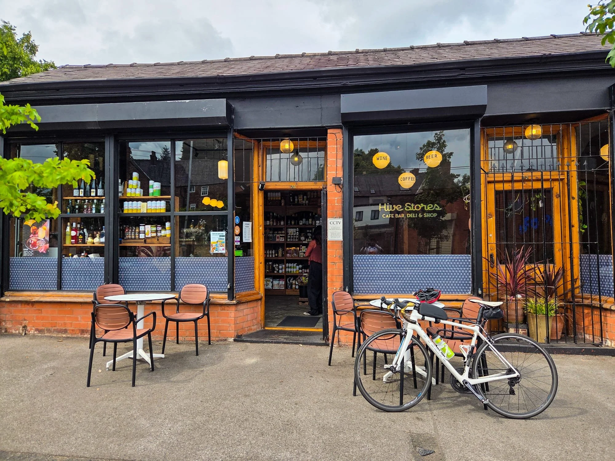 Exterior of Hive Stores, a cafe and shop, with outdoor seating, a bicycle, and signs for wine, beer, coffee, and shop inside.