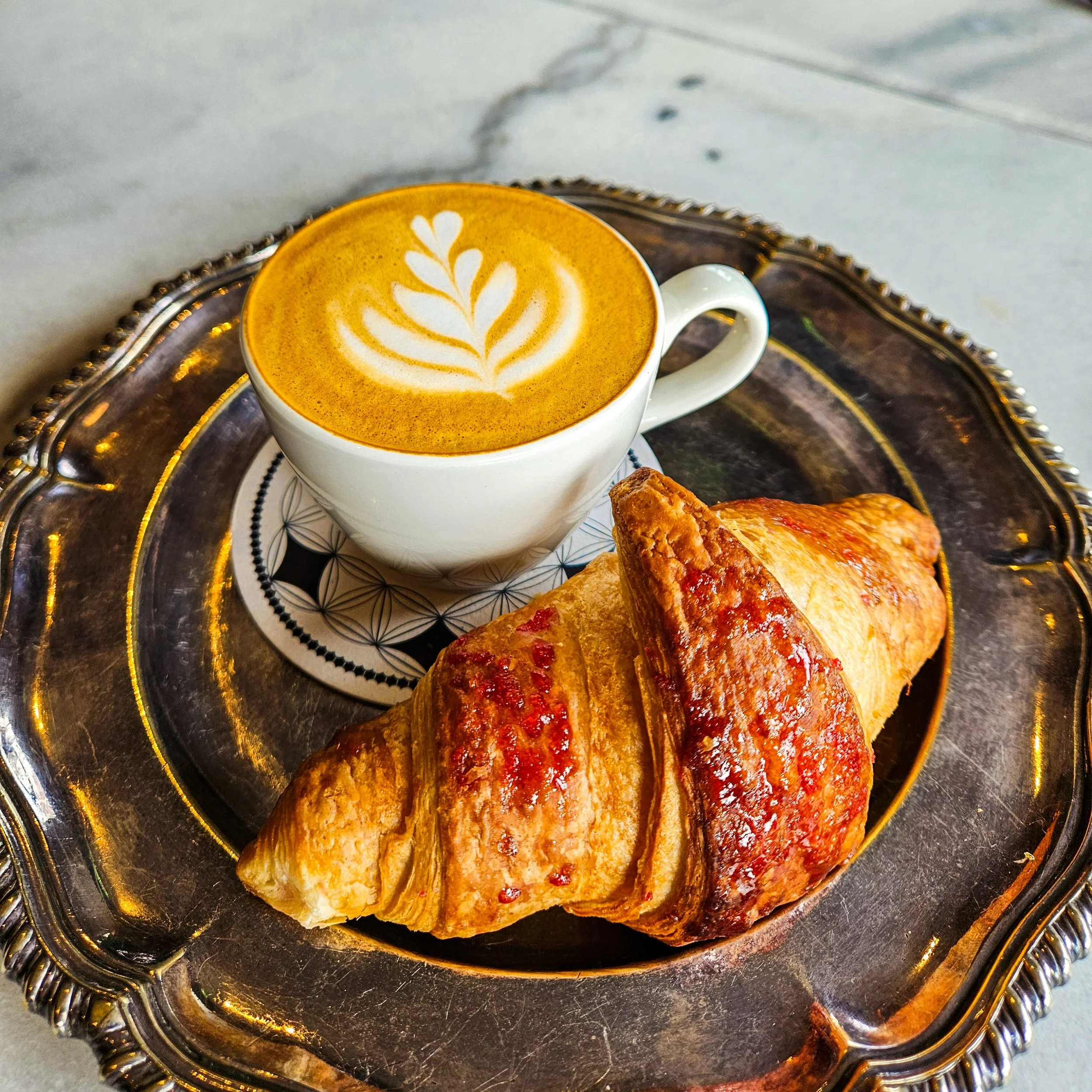 A cup of latte with a leaf-shaped foam art on top, placed on a decorative coaster, along with a croissant on a silver tray.