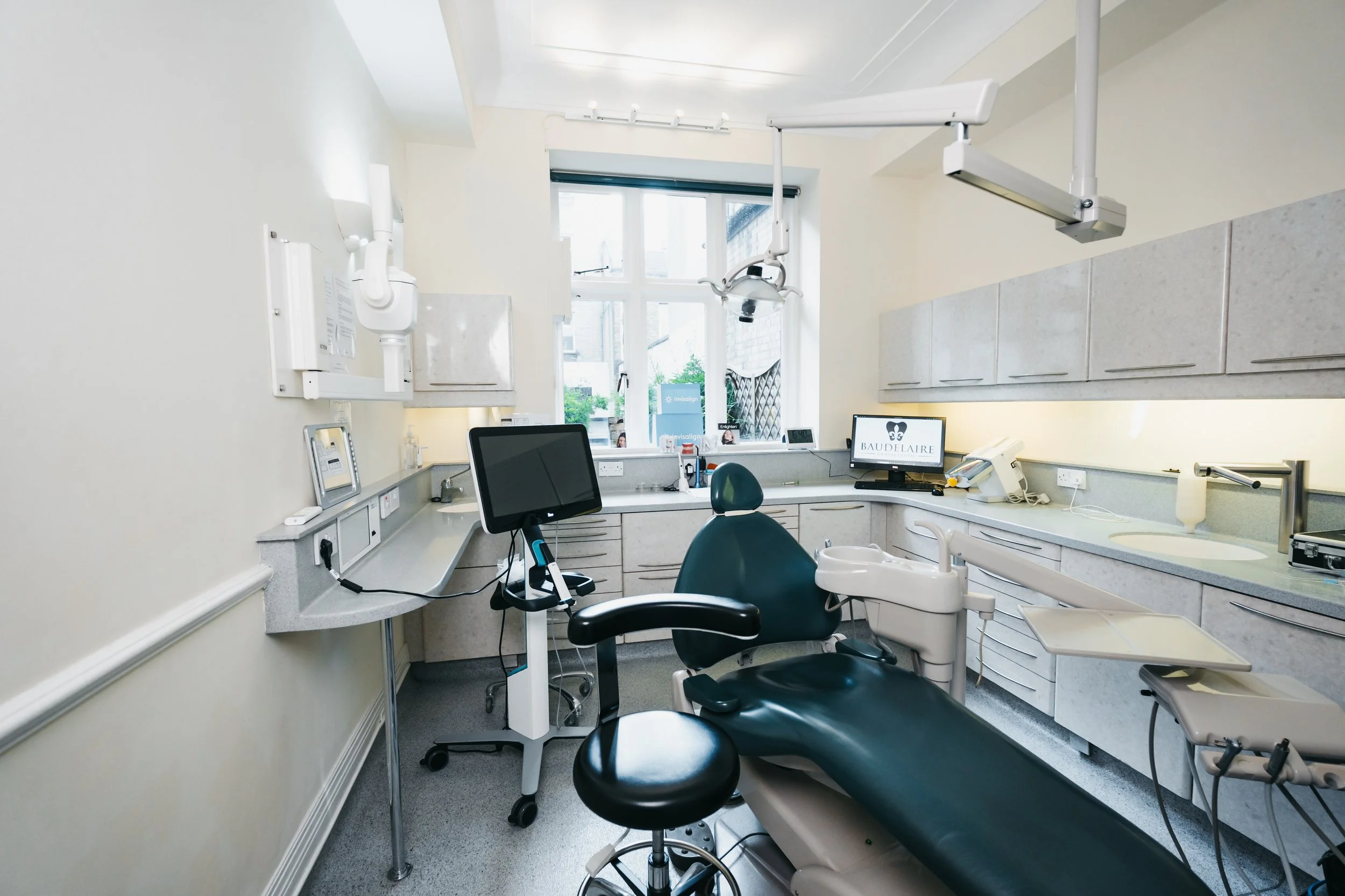 Interior of a dental examination room with a dental chair, monitor screens, dental equipment, and a window with outside views.