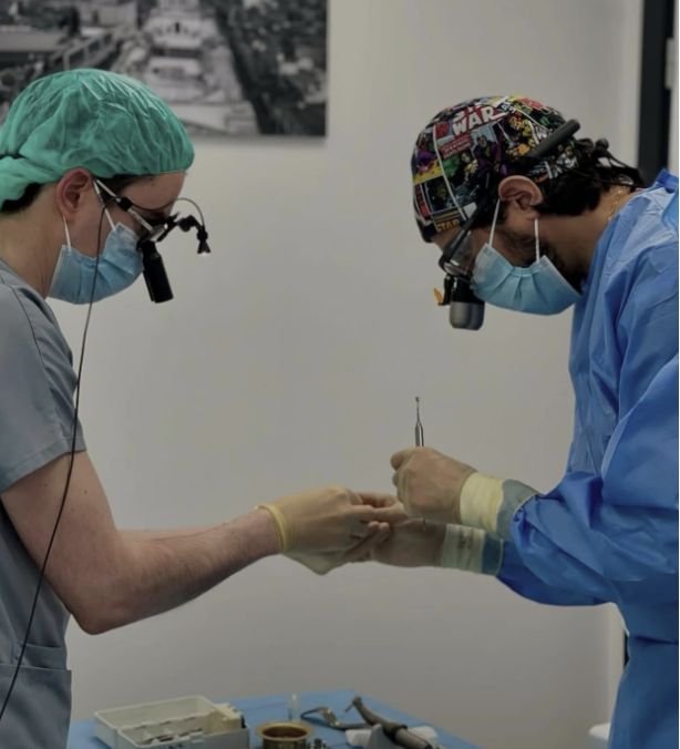 Two surgeons wearing surgical masks, headlamps, and scrubs perform a delicate procedure on a patient's hand, with medical tools on a tray nearby.