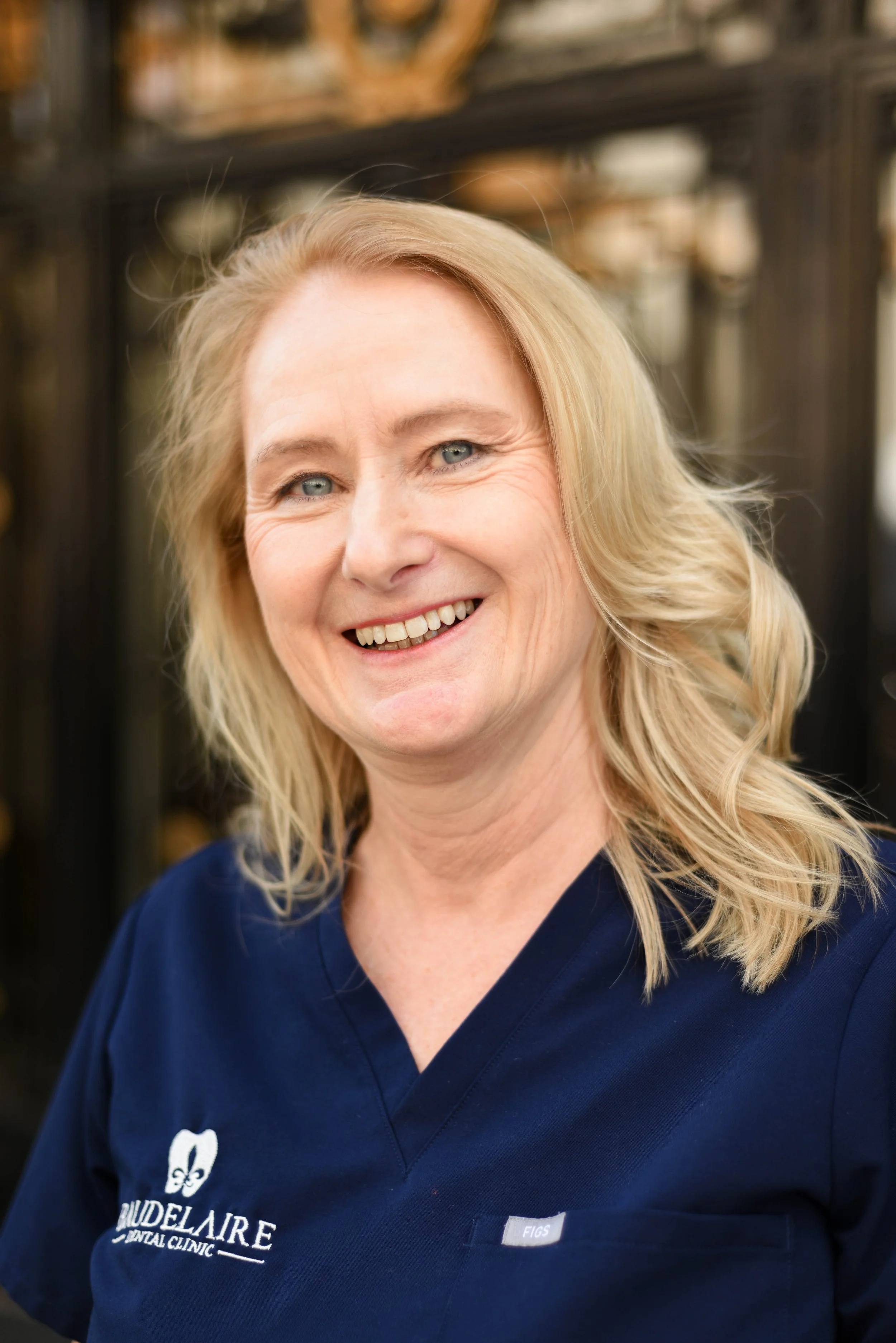 A smiling middle-aged woman with blonde hair wearing a navy blue scrubs with a logo that says "MudelAire Dental Clinic".