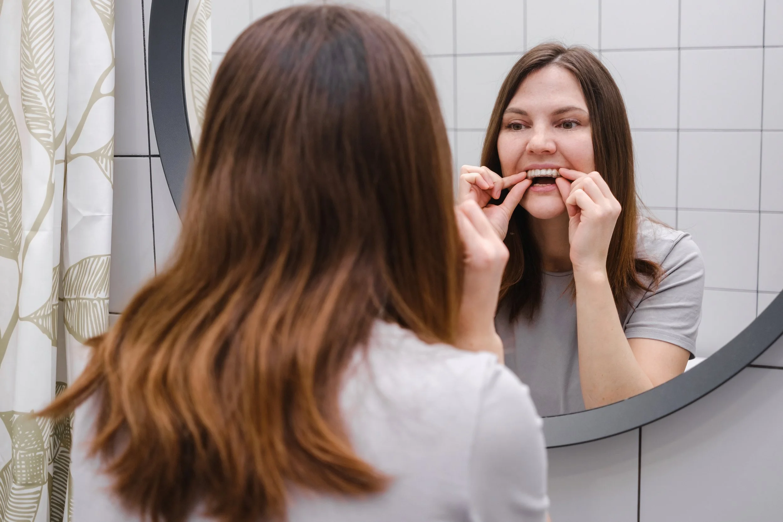 A woman with long brown hair checking her teeth in front of a mirror at a bathroom with white tiled wall.