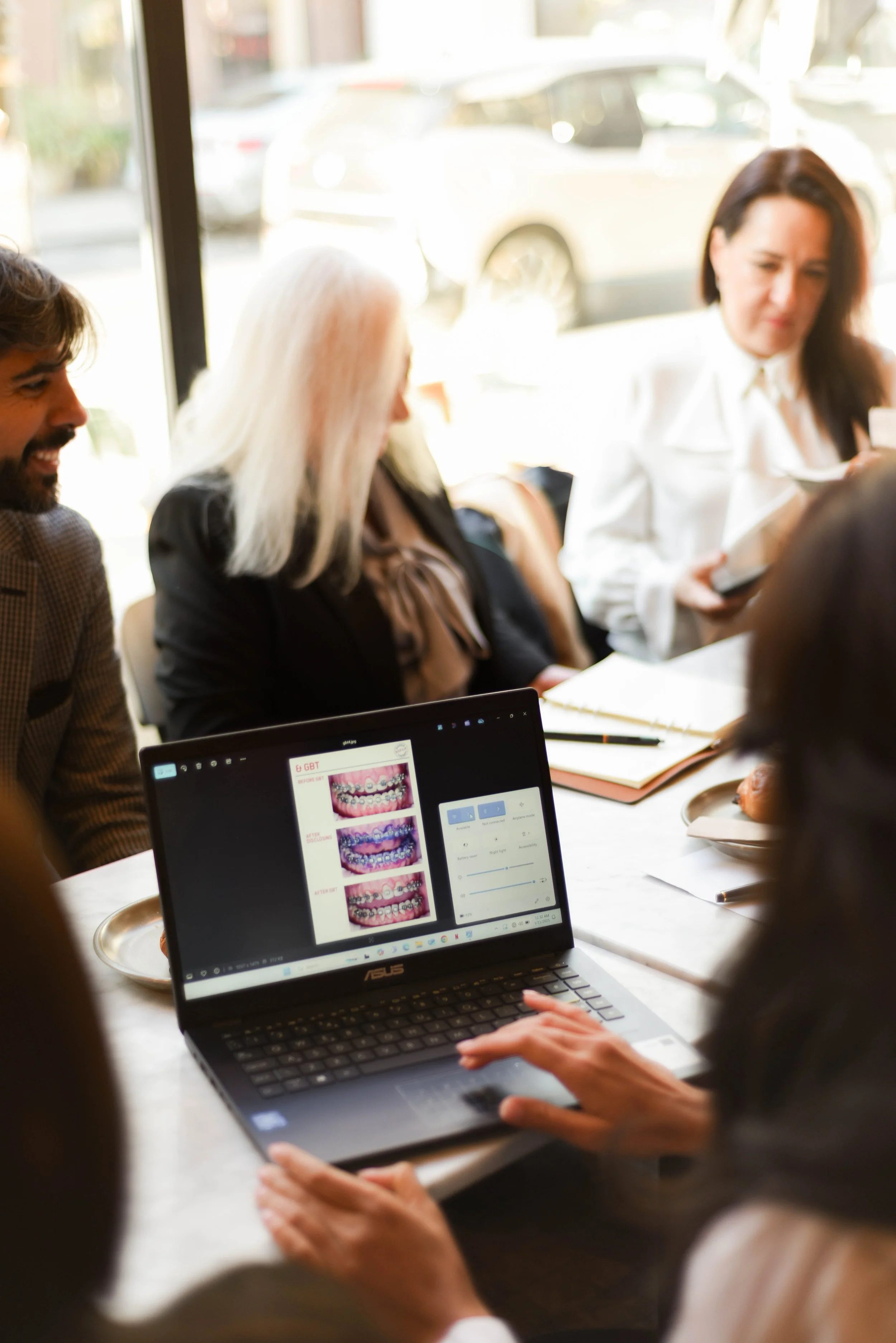 Group of people sitting around a table during a meeting, with a person using a laptop displaying images of teeth and braces.