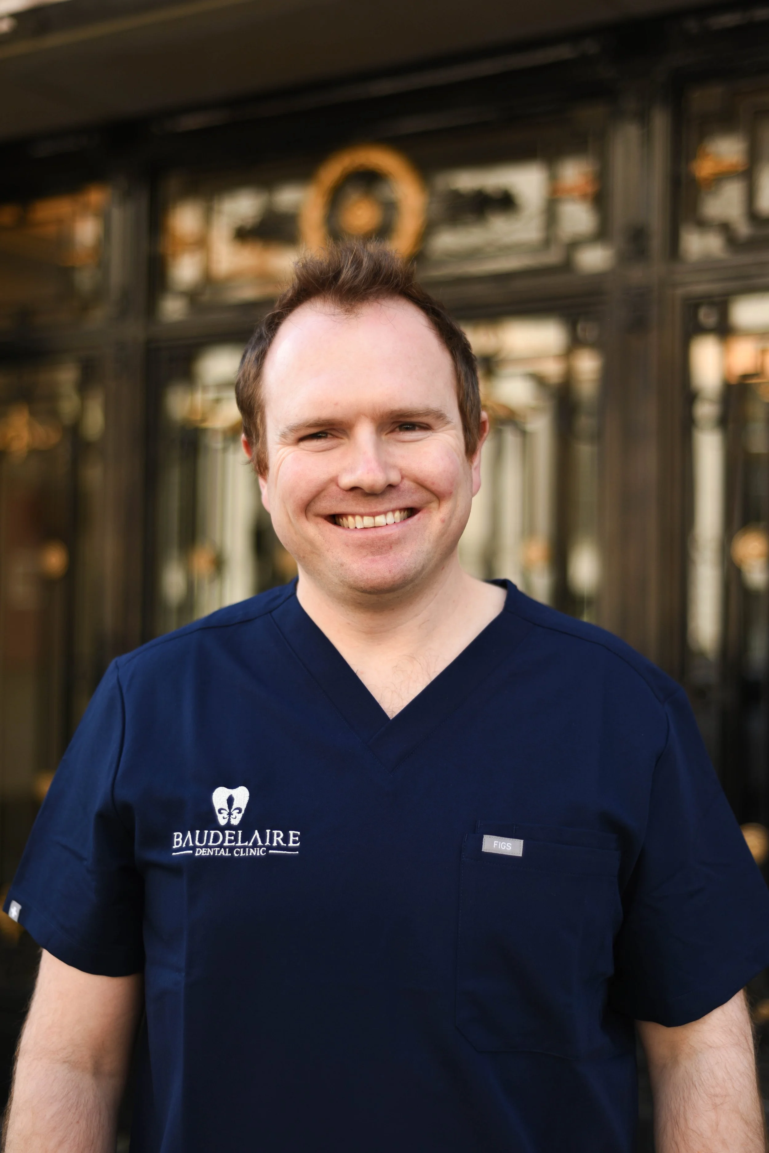 Smiling man in navy dental uniform standing outside in front of a glass building.