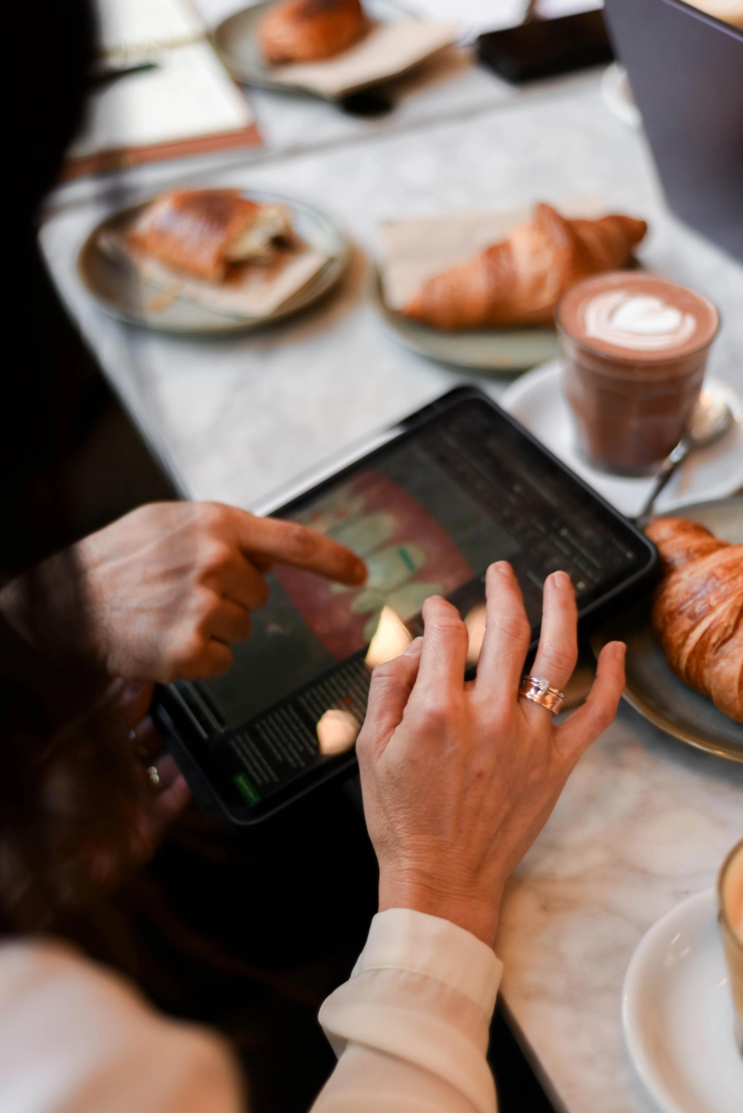 A person is using a tablet to view an image or video at a breakfast table. There are plates with croissants and partially eaten pastries, and a cup of coffee with frothy latte art on a marble table.
