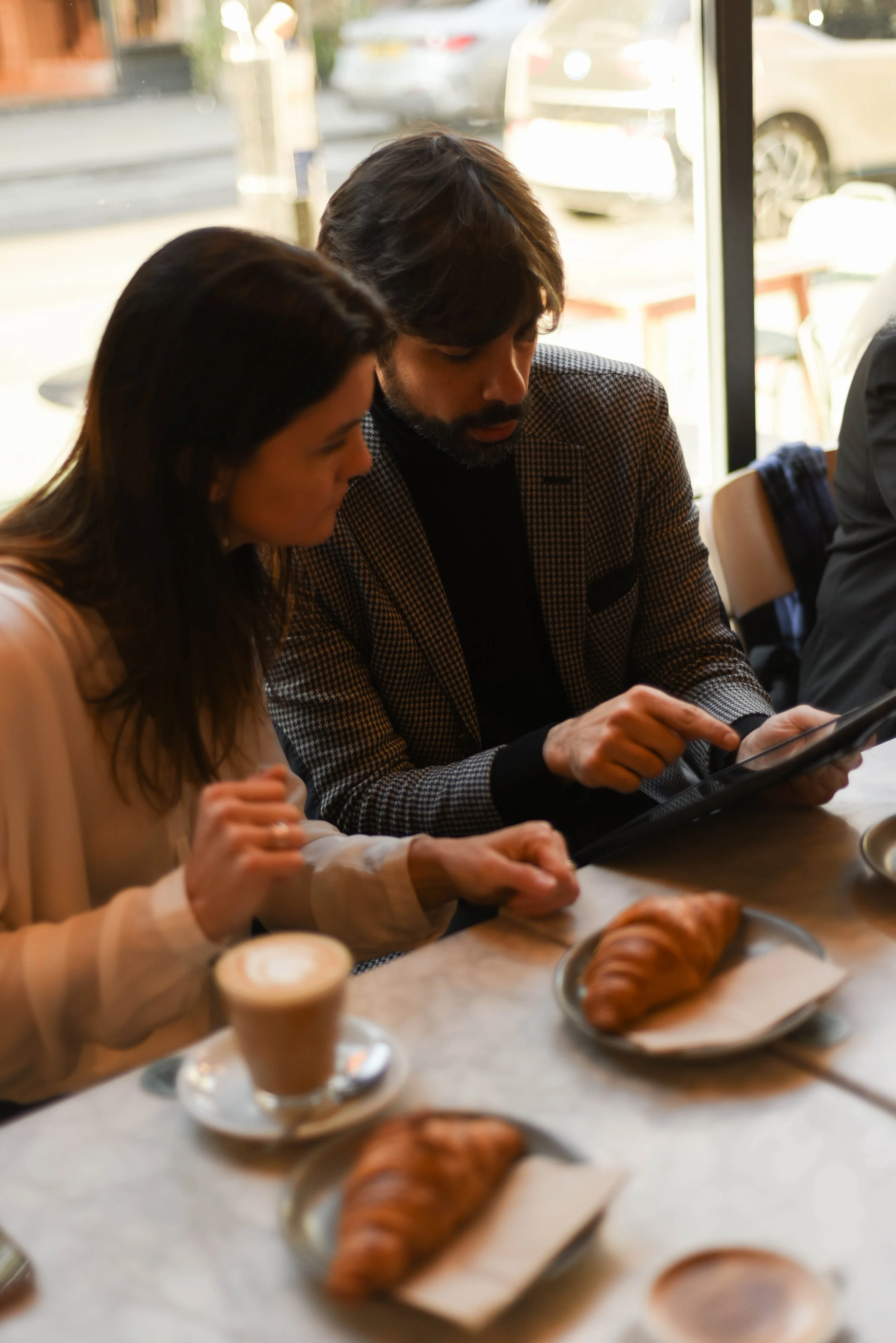 Two people, a woman and a man, sit at a cafe table looking at a tablet together, with croissants and coffee in front of them.