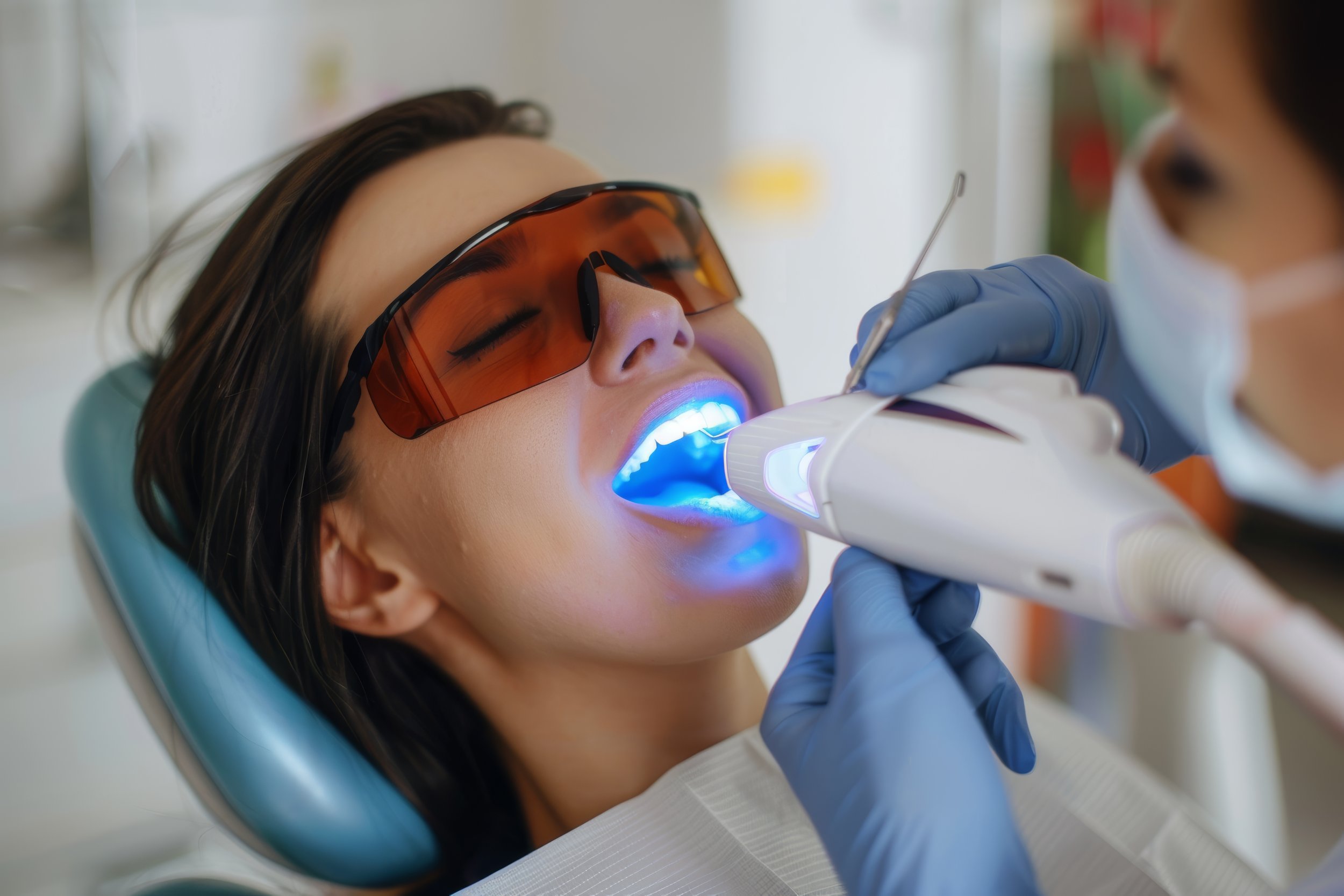 A woman undergoing a dental procedure while wearing protective glasses, with a dental professional using a curing light device in her mouth.