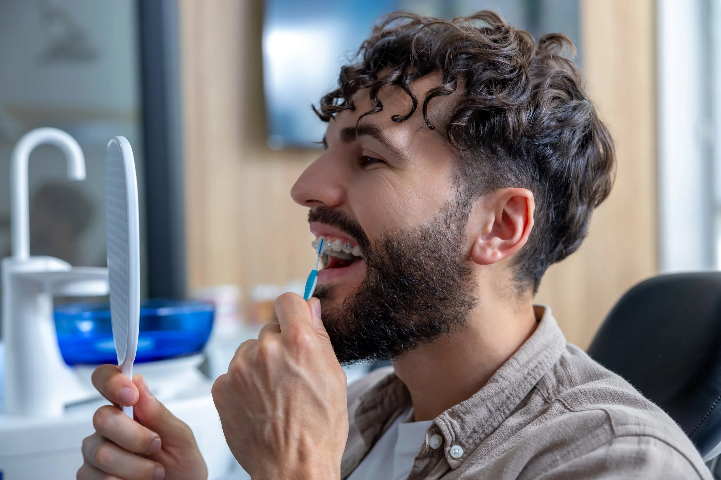 Man with curly hair brushing his teeth while looking at a mirror in a bathroom.
