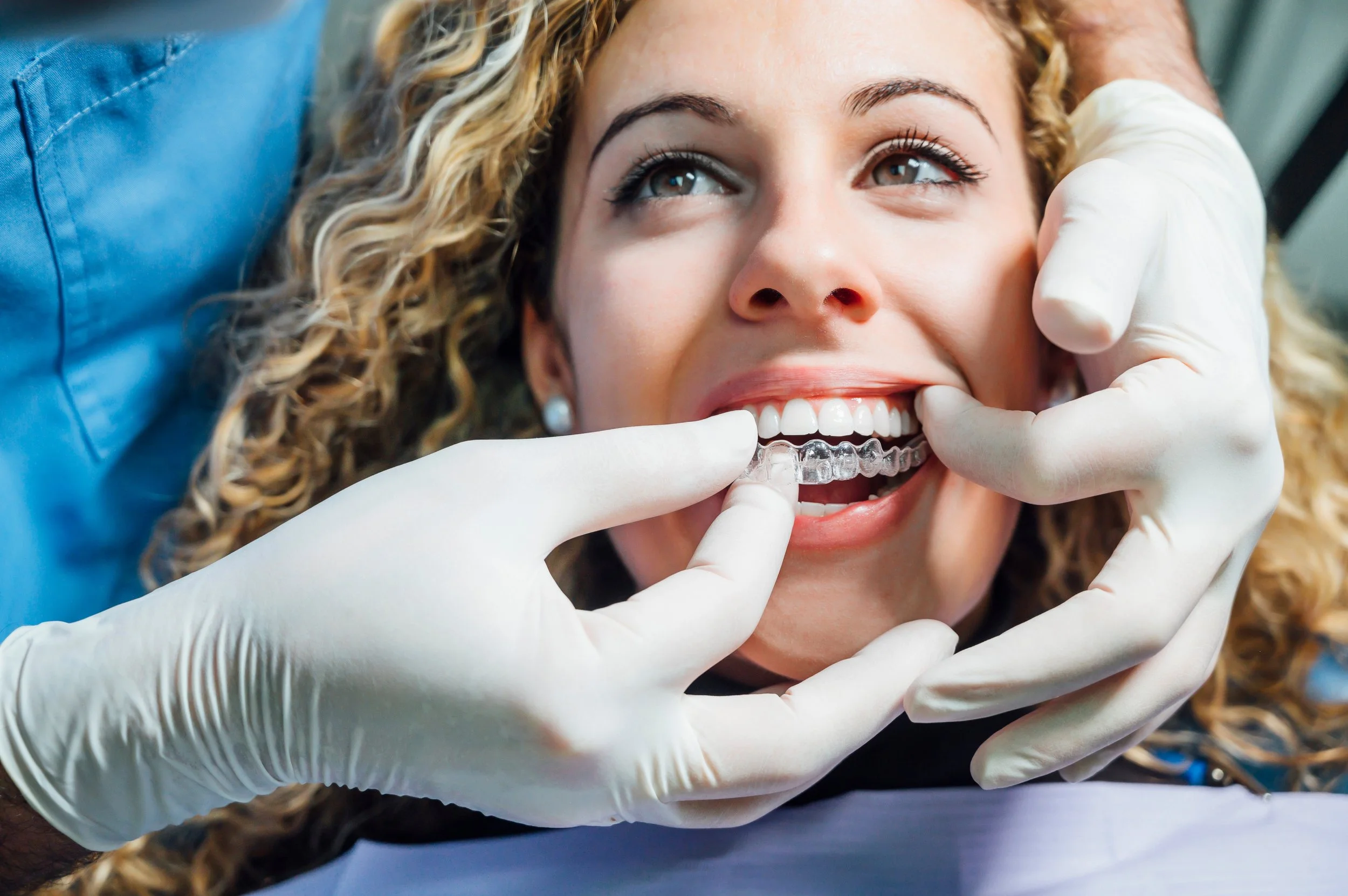 A woman smiling while a dental professional places a clear dental aligner on her teeth.