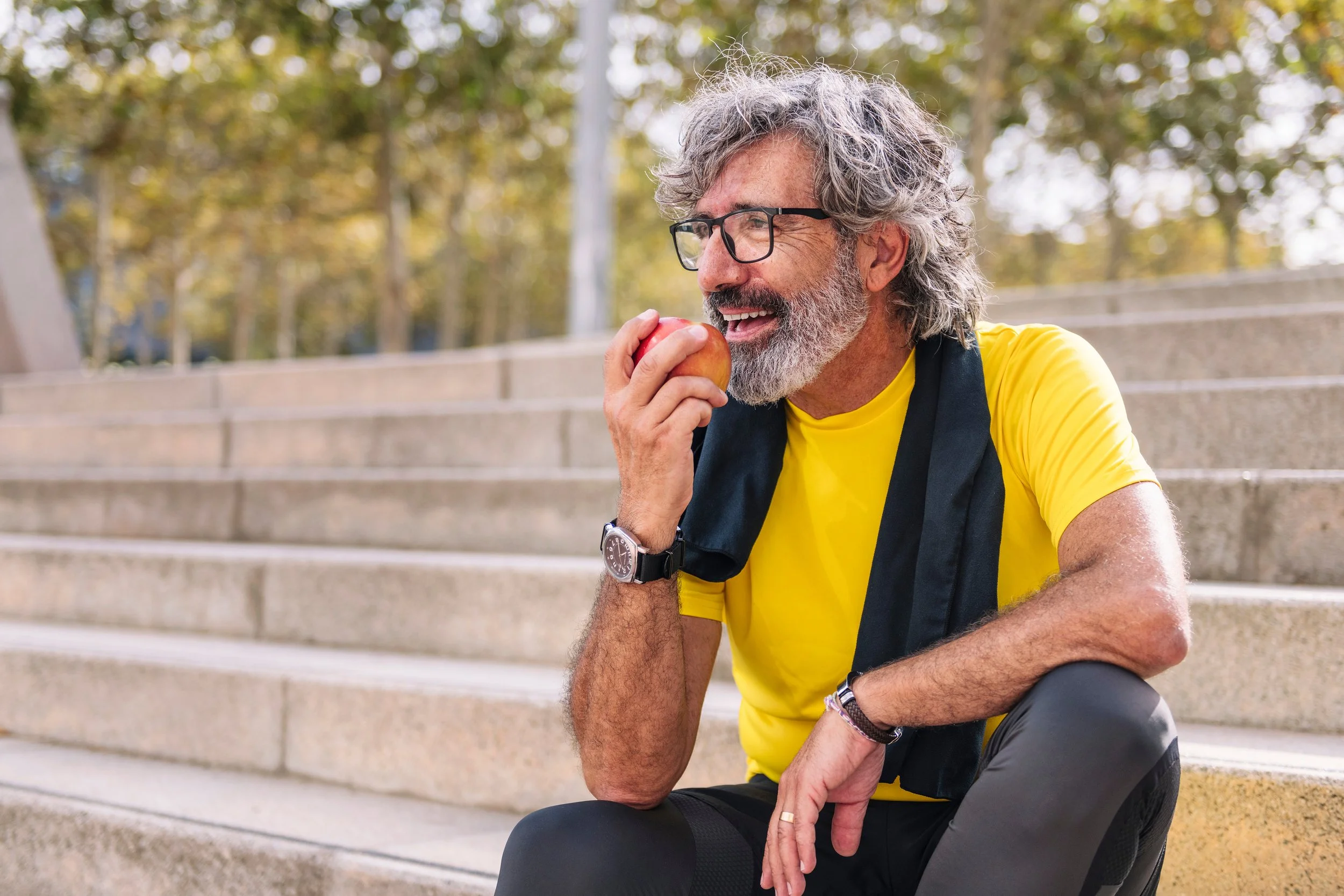 An older man with gray hair and beard, wearing glasses, a yellow T-shirt, and a black vest, smiling and biting into a red apple, sitting outdoors on concrete stairs.