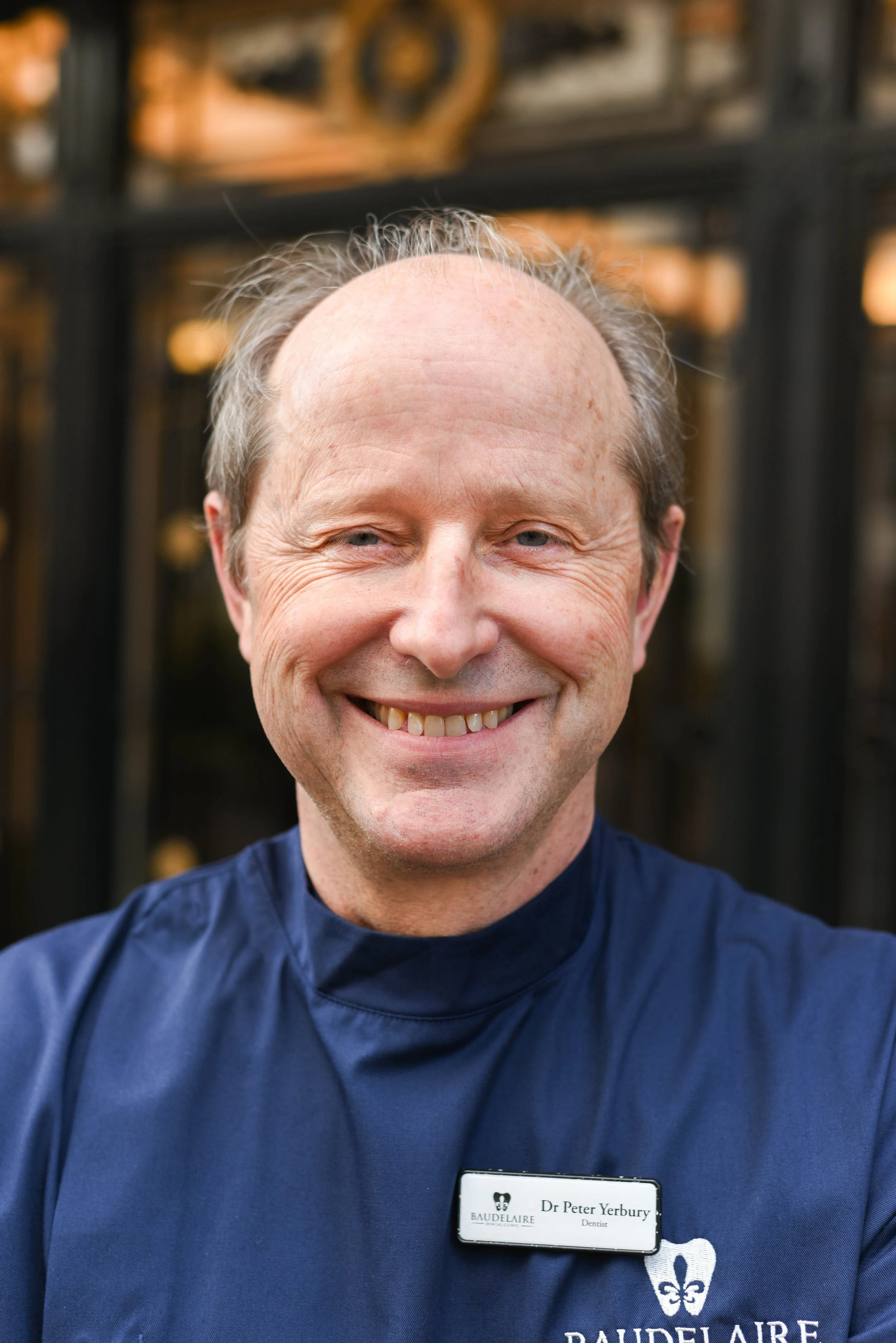 Smiling man wearing a blue dental uniform with a name tag that reads 'Dr. Peter Yerbury, Dentist' in front of a decorative background.