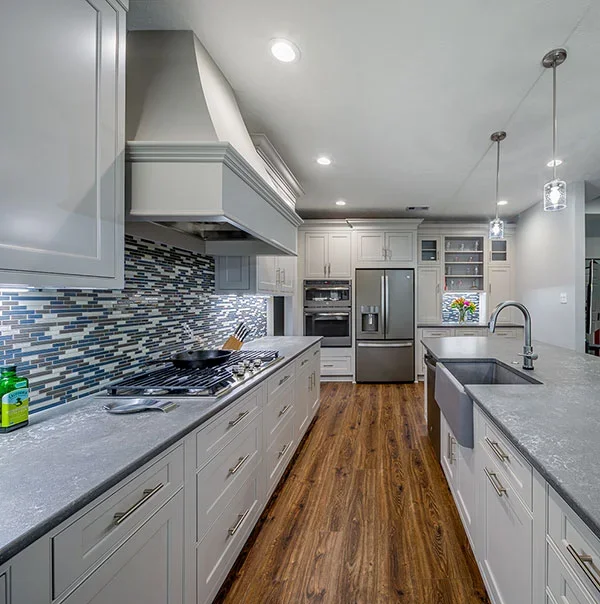 Modern kitchen with white cabinets, gray countertops, a stainless steel refrigerator, and a mosaic tile backsplash; wood flooring and pendant lights over the island.
