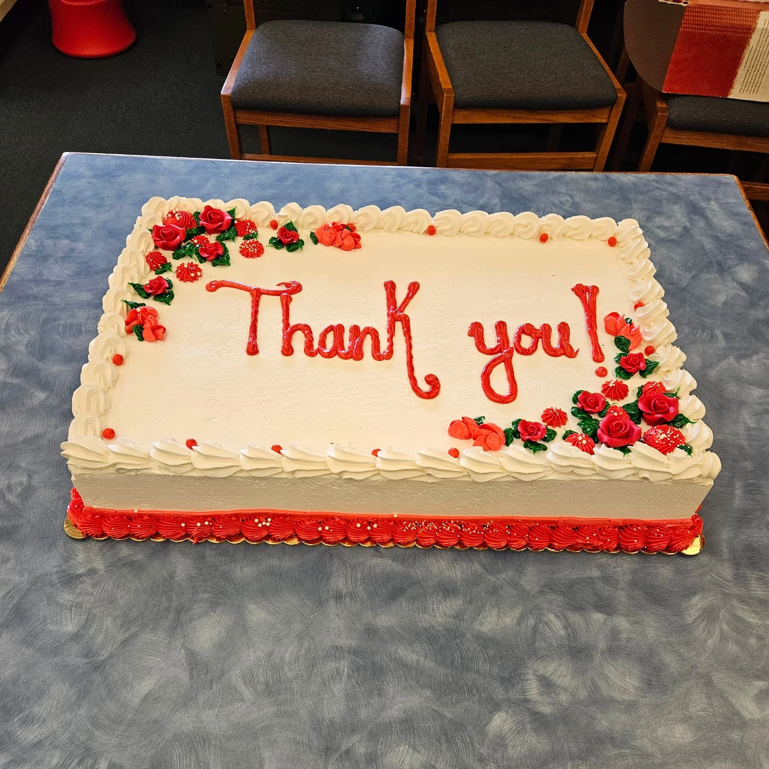 A rectangular birthday cake with white icing, decorated with red and pink flowers made of frosting at the top left and bottom right corners. The cake has a red border along the bottom edge and the words "Thank you!" written in red icing in the center
