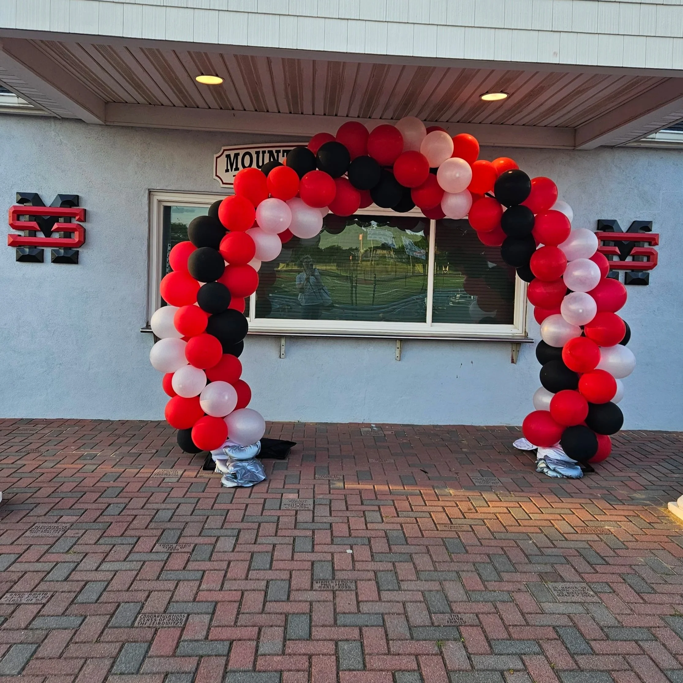 Balloon arch with red, black, and white balloons in front of a white building with a window.