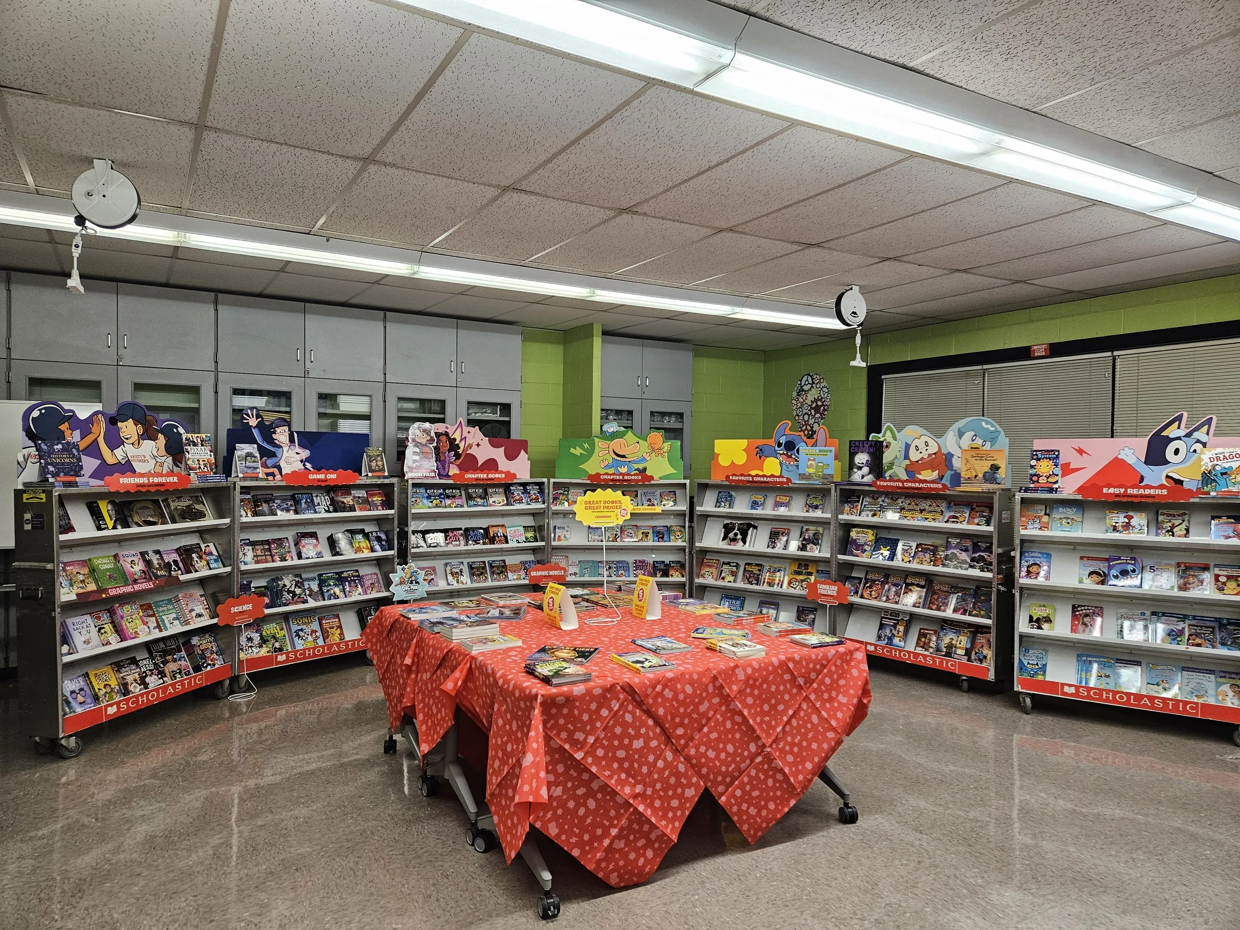 Kids' book display in a school library with a red tablecloth and shelves filled with colorful books and cartoon character decorations.