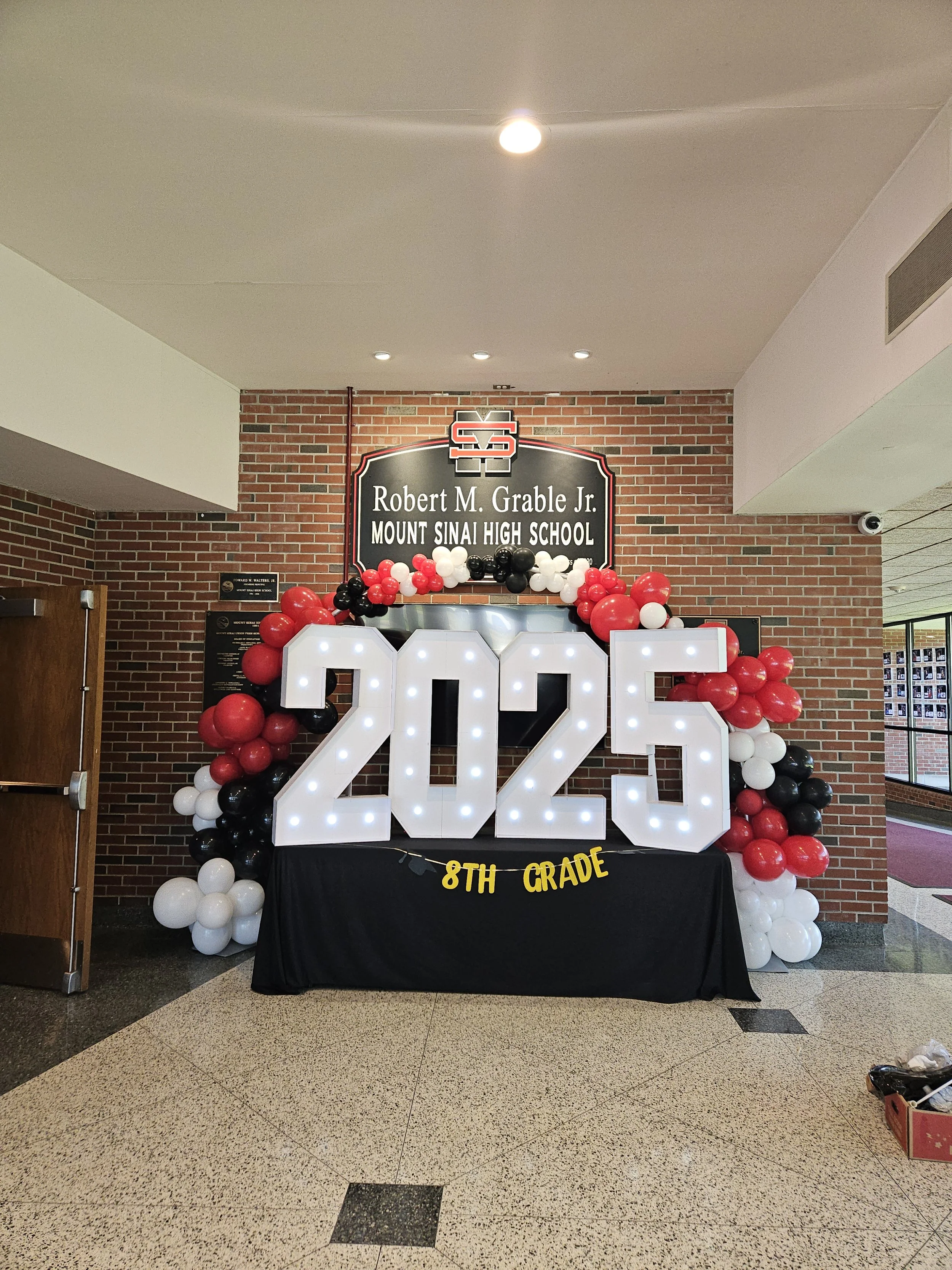Decorative display celebrating the 2025 8th grade graduation at Robert M. Grable Jr. Mount Sinai High School, featuring large illuminated numbers '2025,' balloons in black, white, and red, and a black table with '8th Grade' banner.
