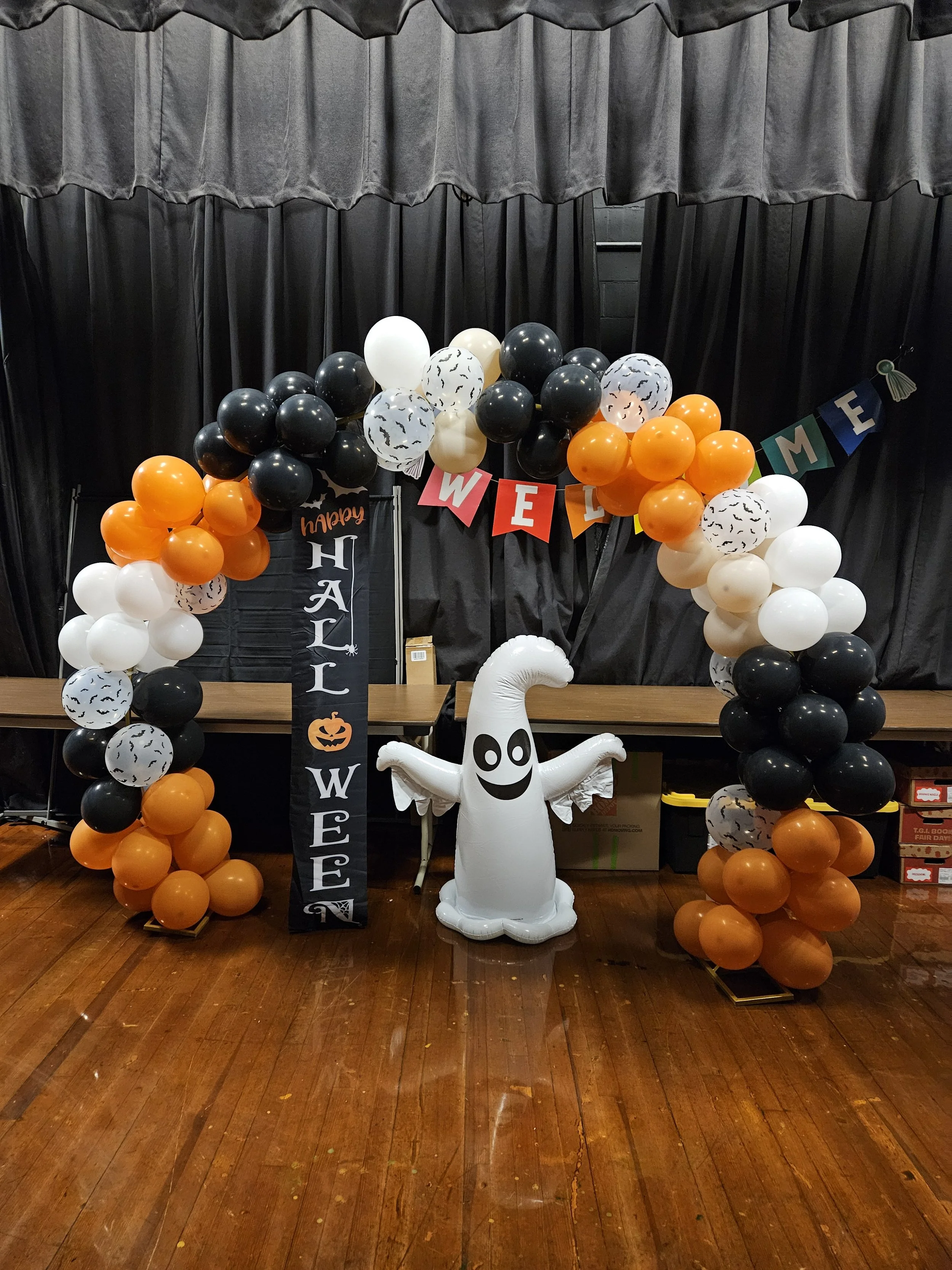 Halloween party decoration with black, white, orange balloons forming an arch. A white ghost-shaped balloon with a smiling face stands in front. A black vertical banner with