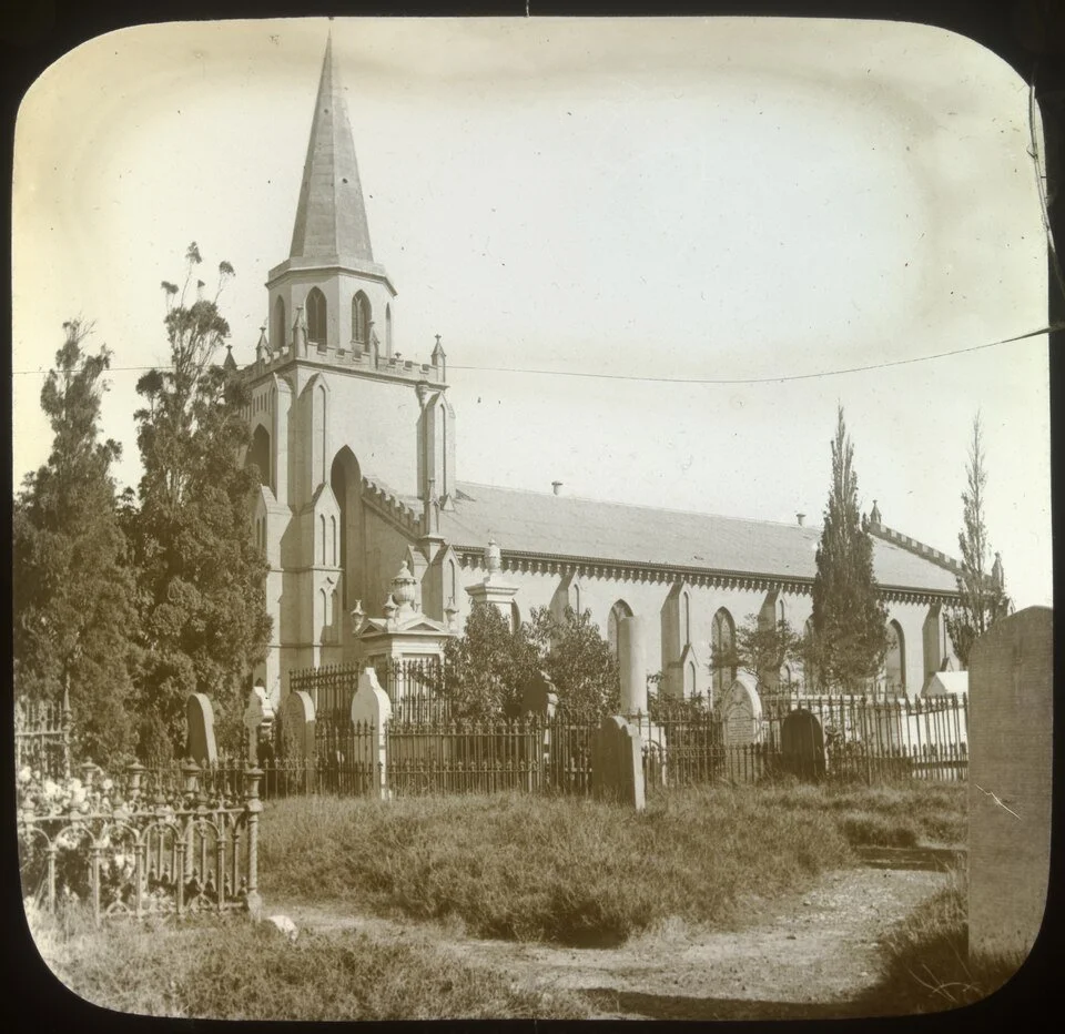 An old black and white photograph of a large church with a tall steeple, surrounded by a wrought iron fence and trees, with a dirt path in the foreground.