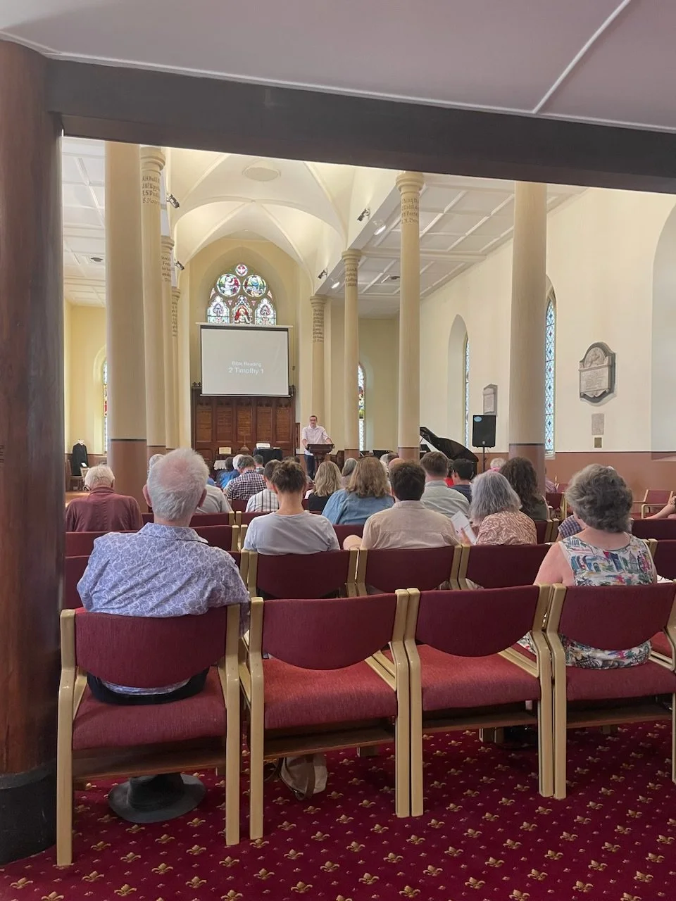 People sitting in a church during a service, with a speaker at the pulpit and a projection screen behind them. The church has tall columns, stained glass windows, and a red patterned carpet.
