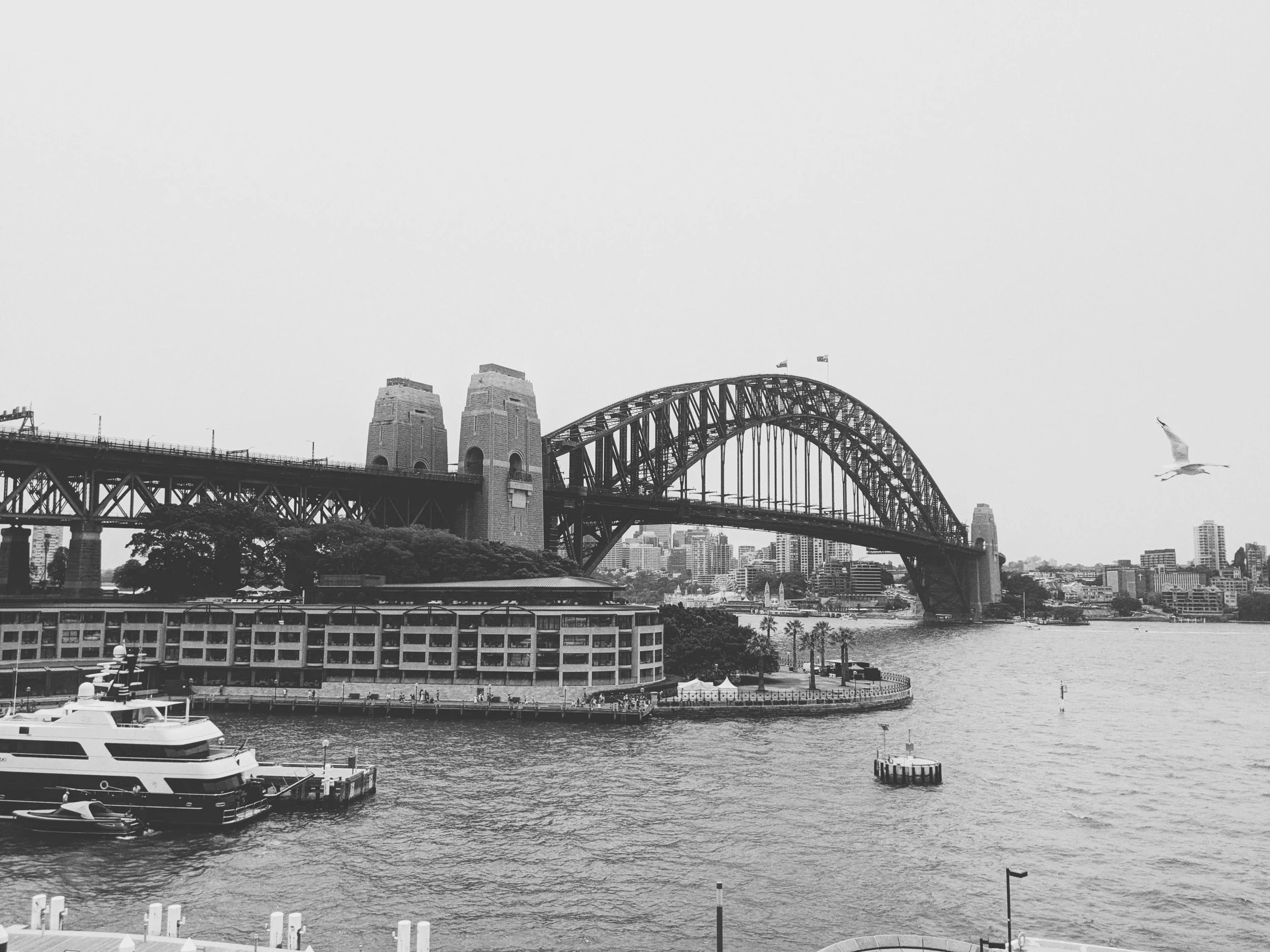 A black and white photo of a cityscape with a large arched bridge spanning a river, with buildings in the background and boats in the water.