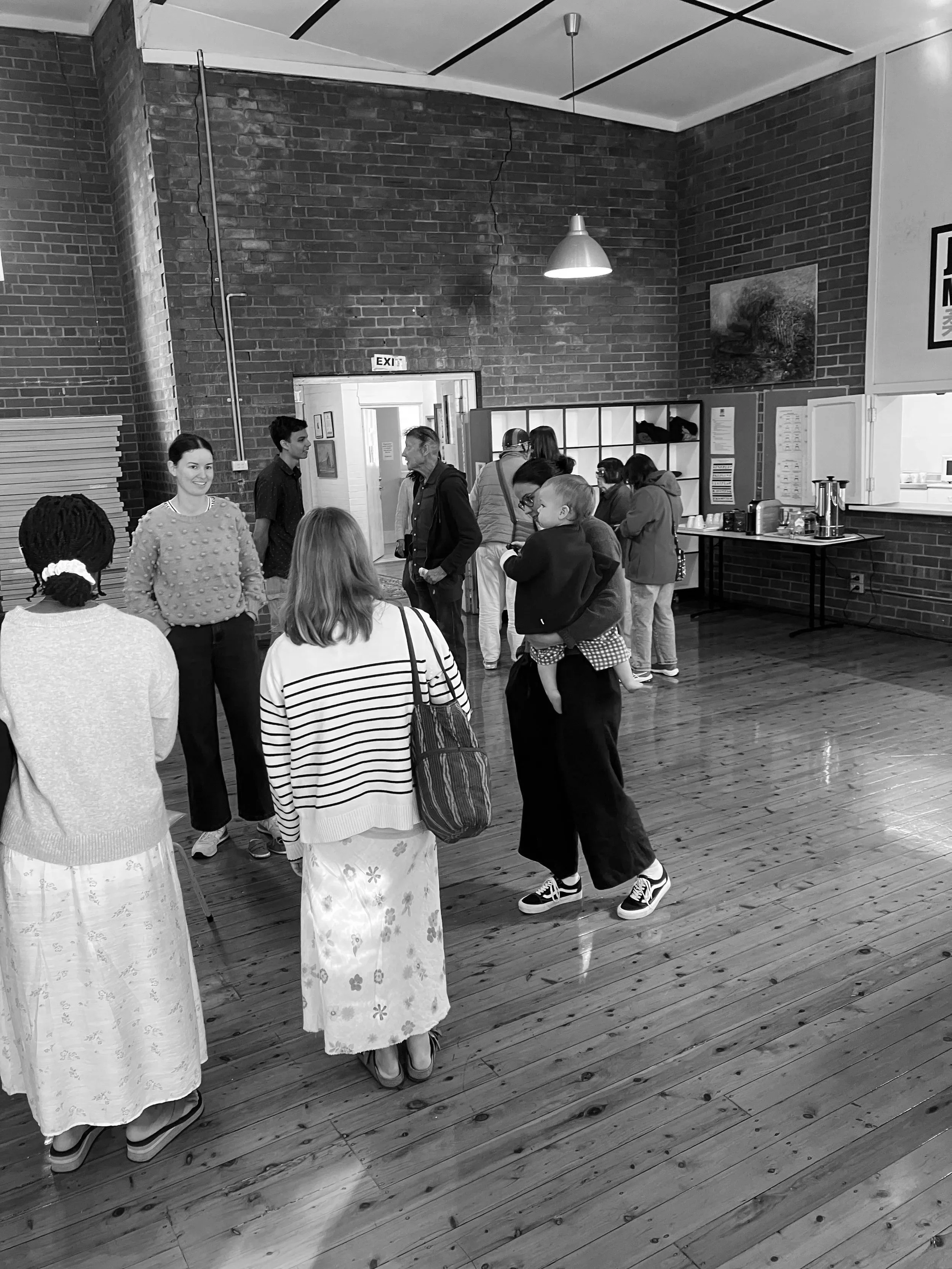 A group of people in a room with exposed brick walls and hardwood floors, some dressed in traditional clothing, others in casual attire, standing and conversing near a table with coffee supplies, with a sign labeled 'EXIT' above a door in the background.