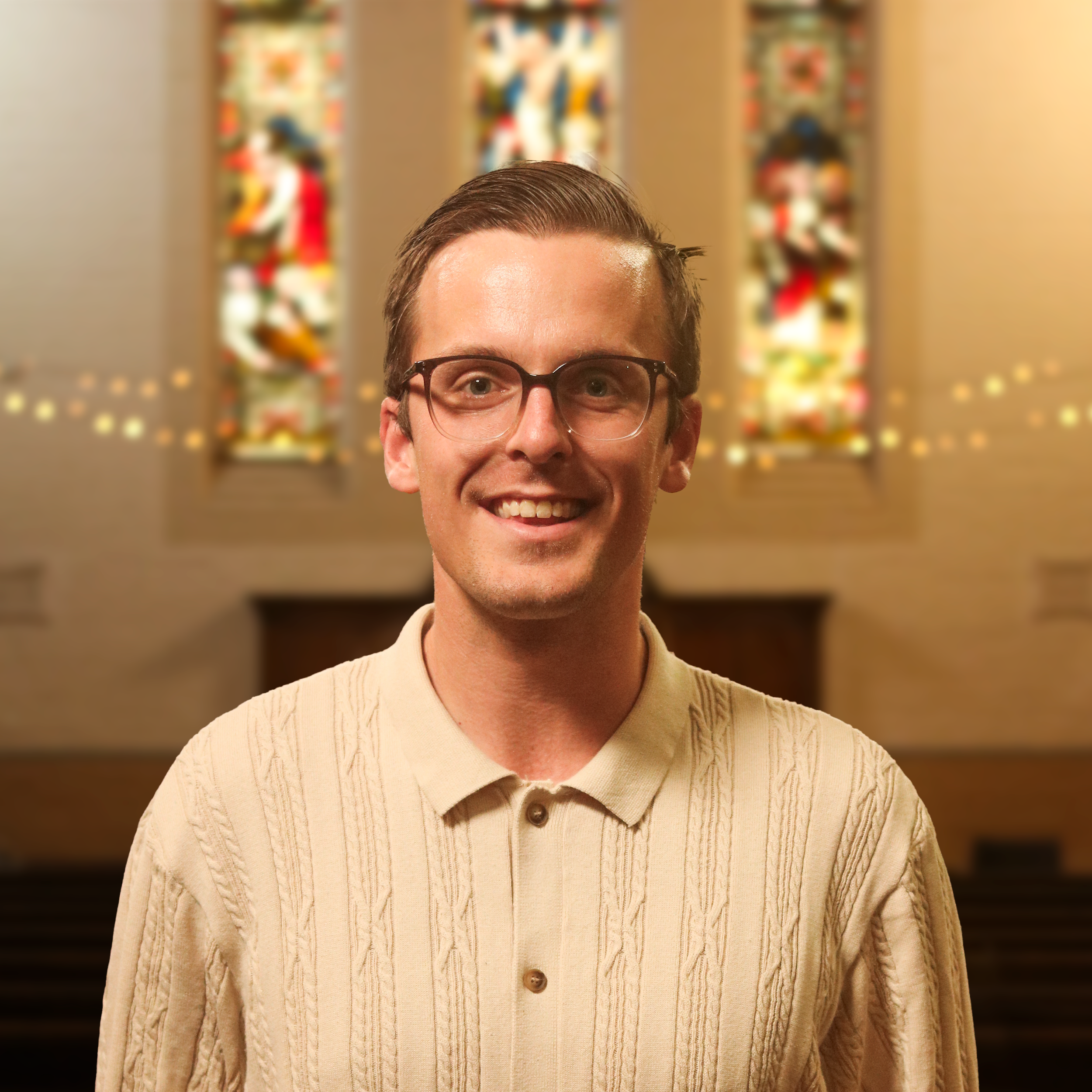 Close-up of a smiling man with glasses, wearing a beige sweater, standing in front of colorful vertical artworks.