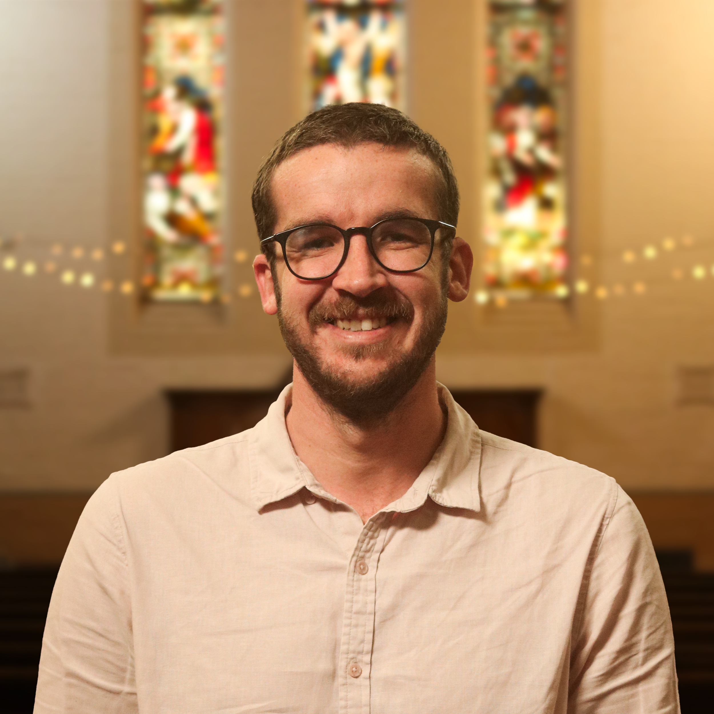 A smiling man with glasses and a light-colored shirt standing in front of blurred colorful artwork or stained glass panels.