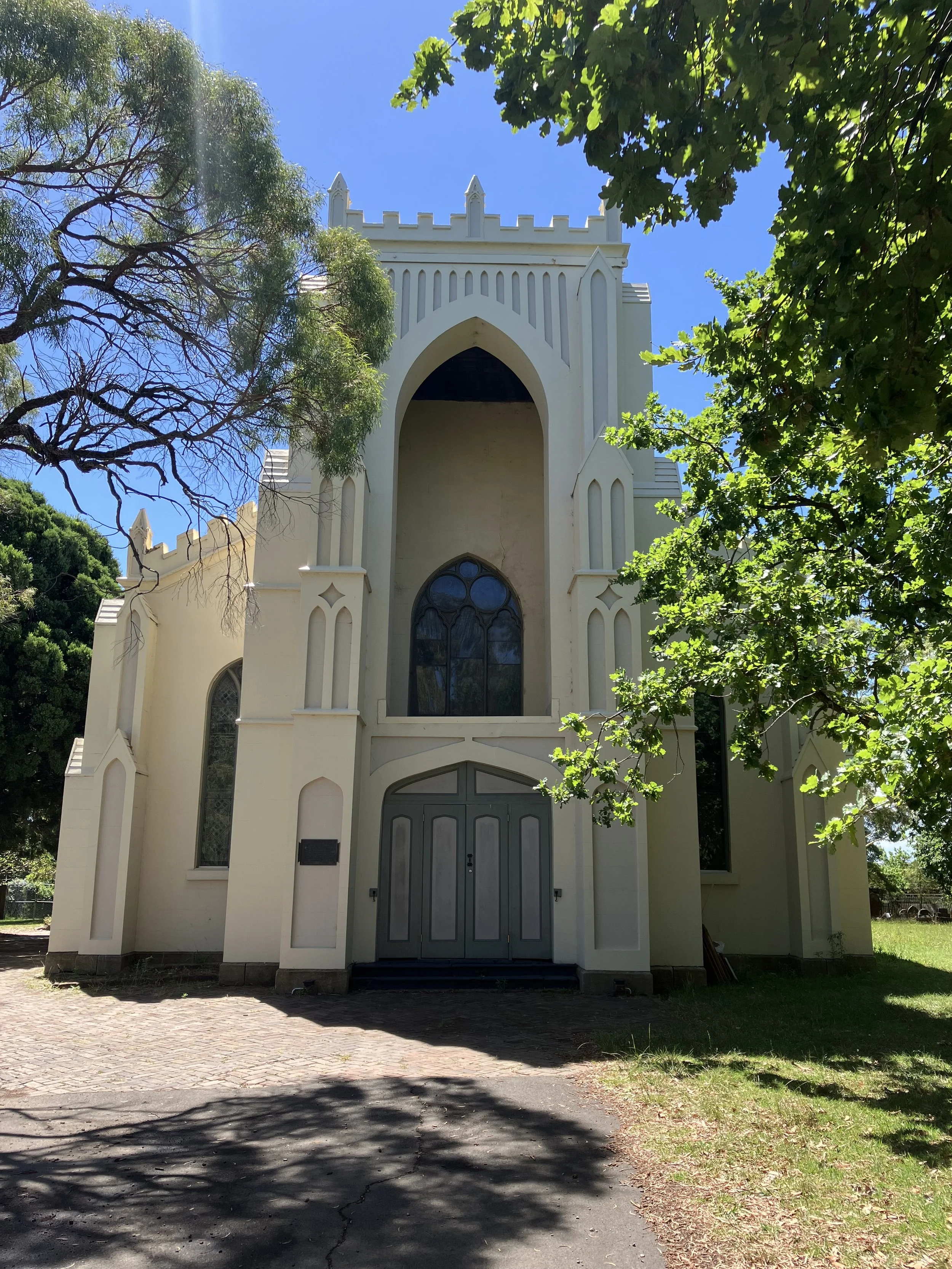A white church with pointed arches and crenellations at the top, surrounded by green trees and under a clear blue sky.