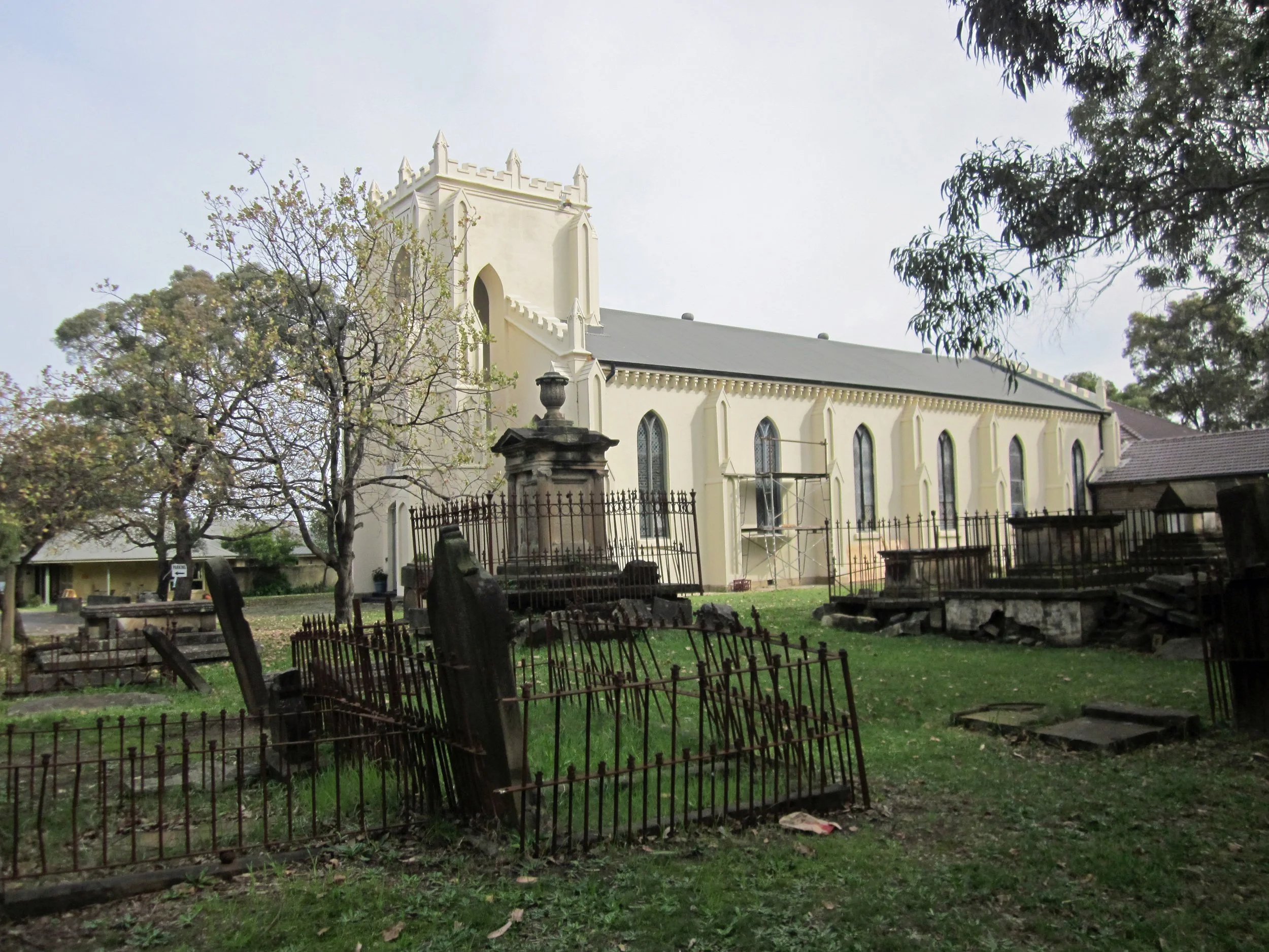 A white church building with gothic windows and a gray roof, surrounded by leafless and leafy trees, in a graveyard with old, tilted, metal fences and gravestones.