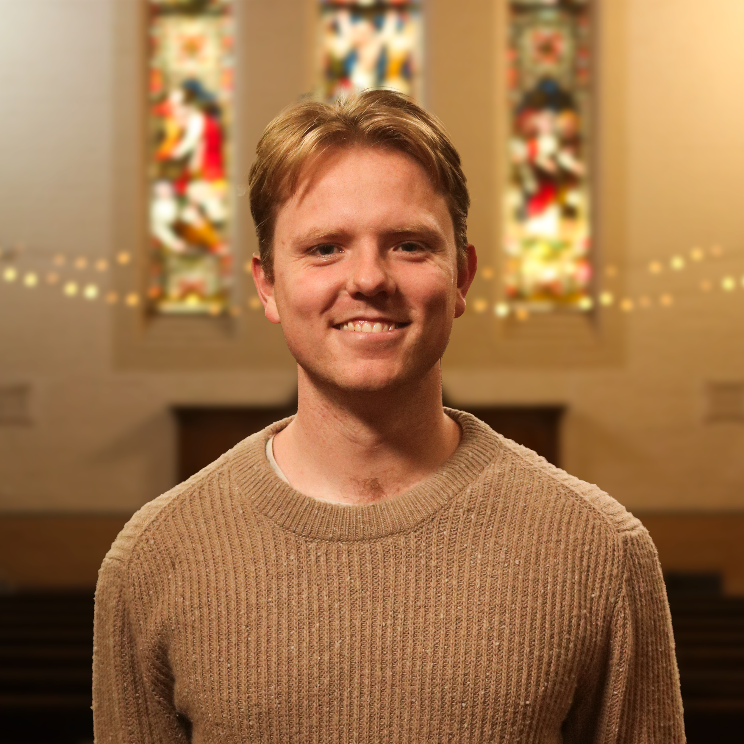 Close-up of a young man with light brown hair, smiling, wearing a beige knitted sweater, with colorful abstract artwork in the background.